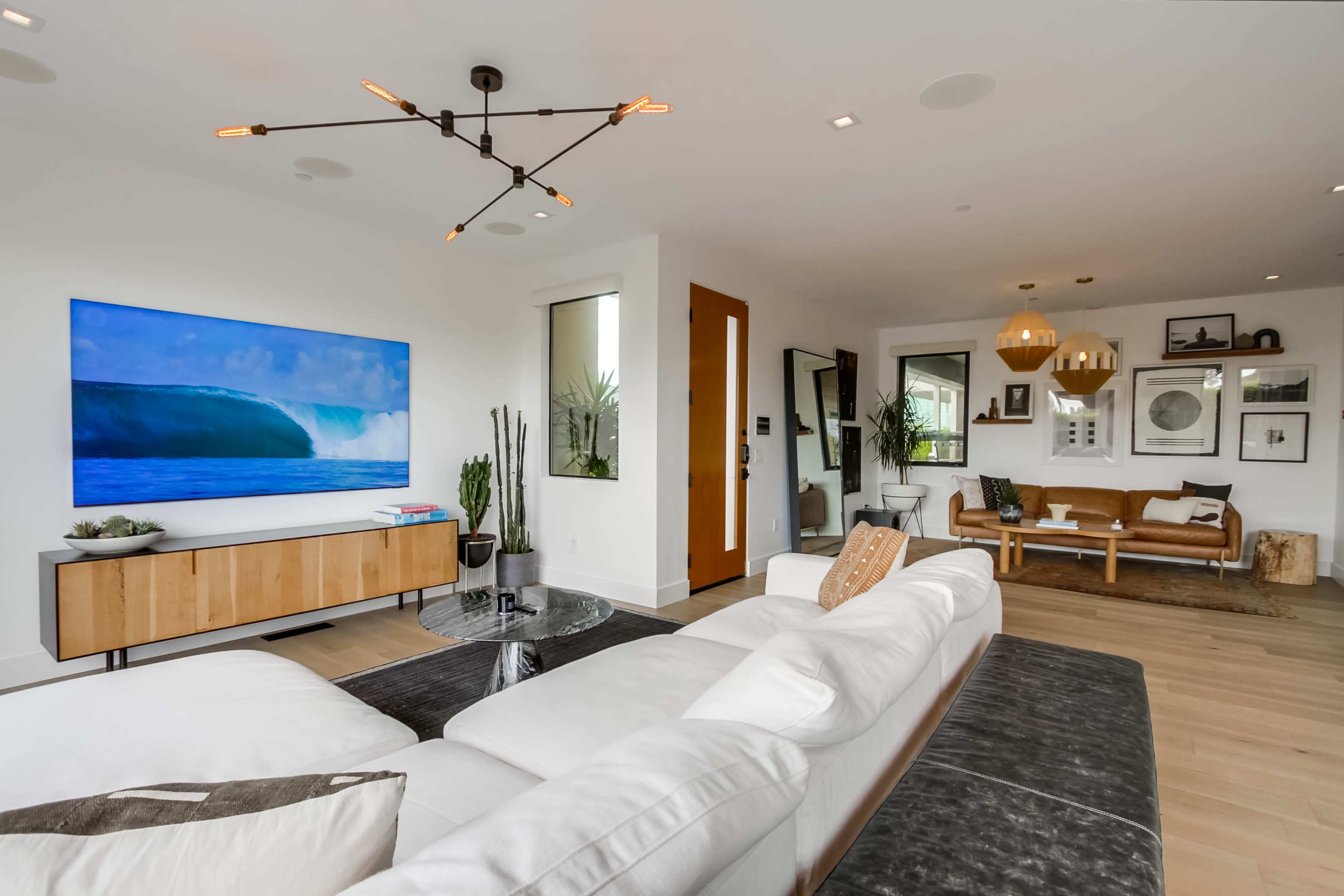A modern living room features a large television on a light wood entertainment unit, a sectional sofa, and decorative plants, complemented by stylish light fixtures and artwork on the walls.