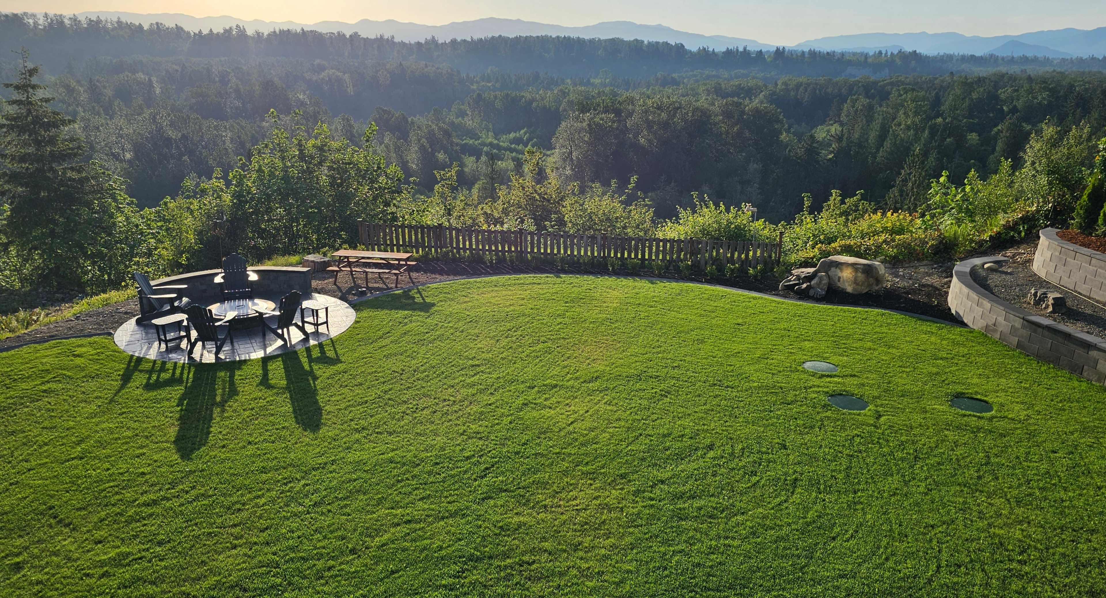 The image shows a well-maintained green lawn with outdoor seating arranged in a circular pattern and a scenic view of rolling hills in the background.