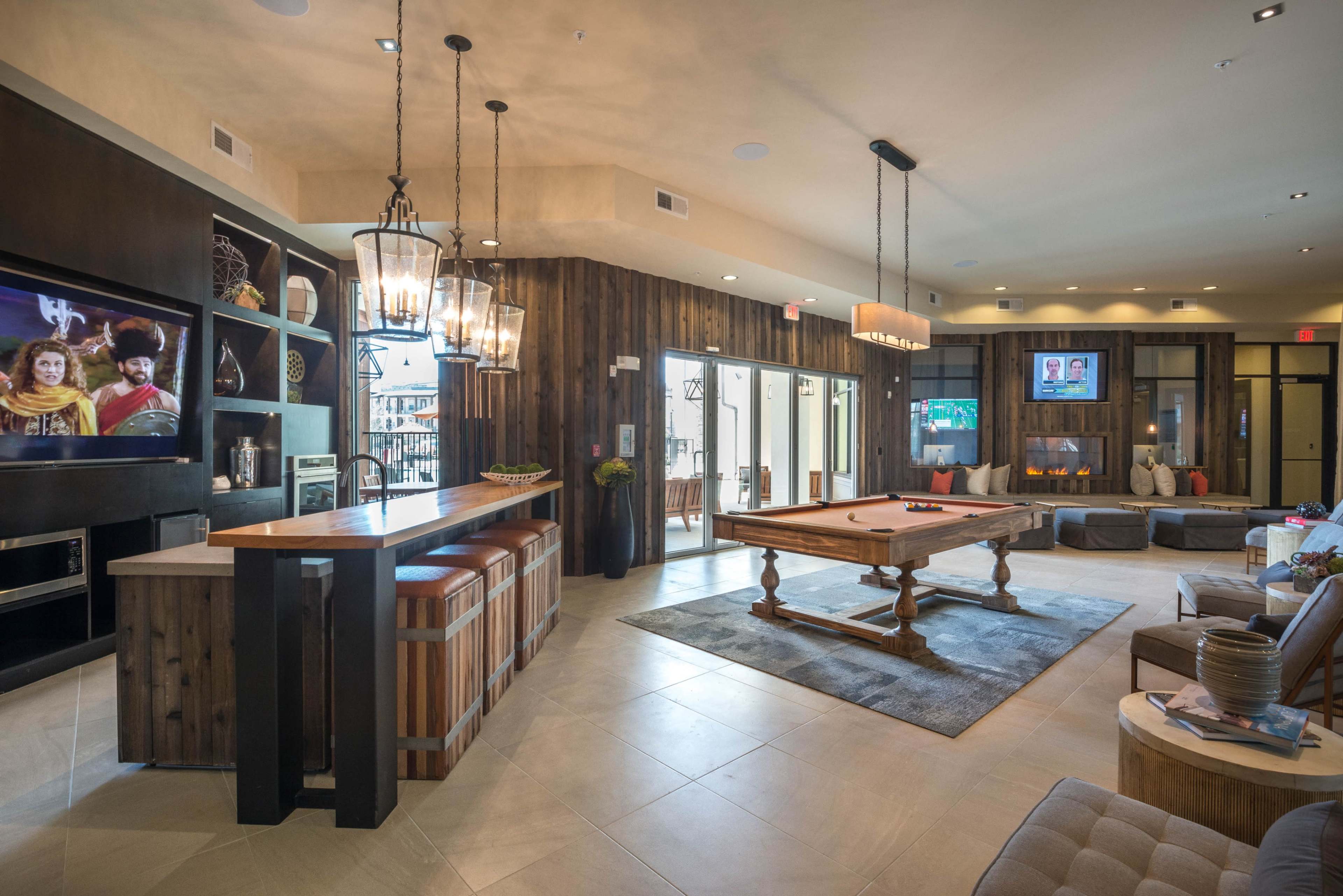 The image depicts a modern lounge area featuring a bar, pool table, and comfortable seating, with large windows allowing natural light to enter.