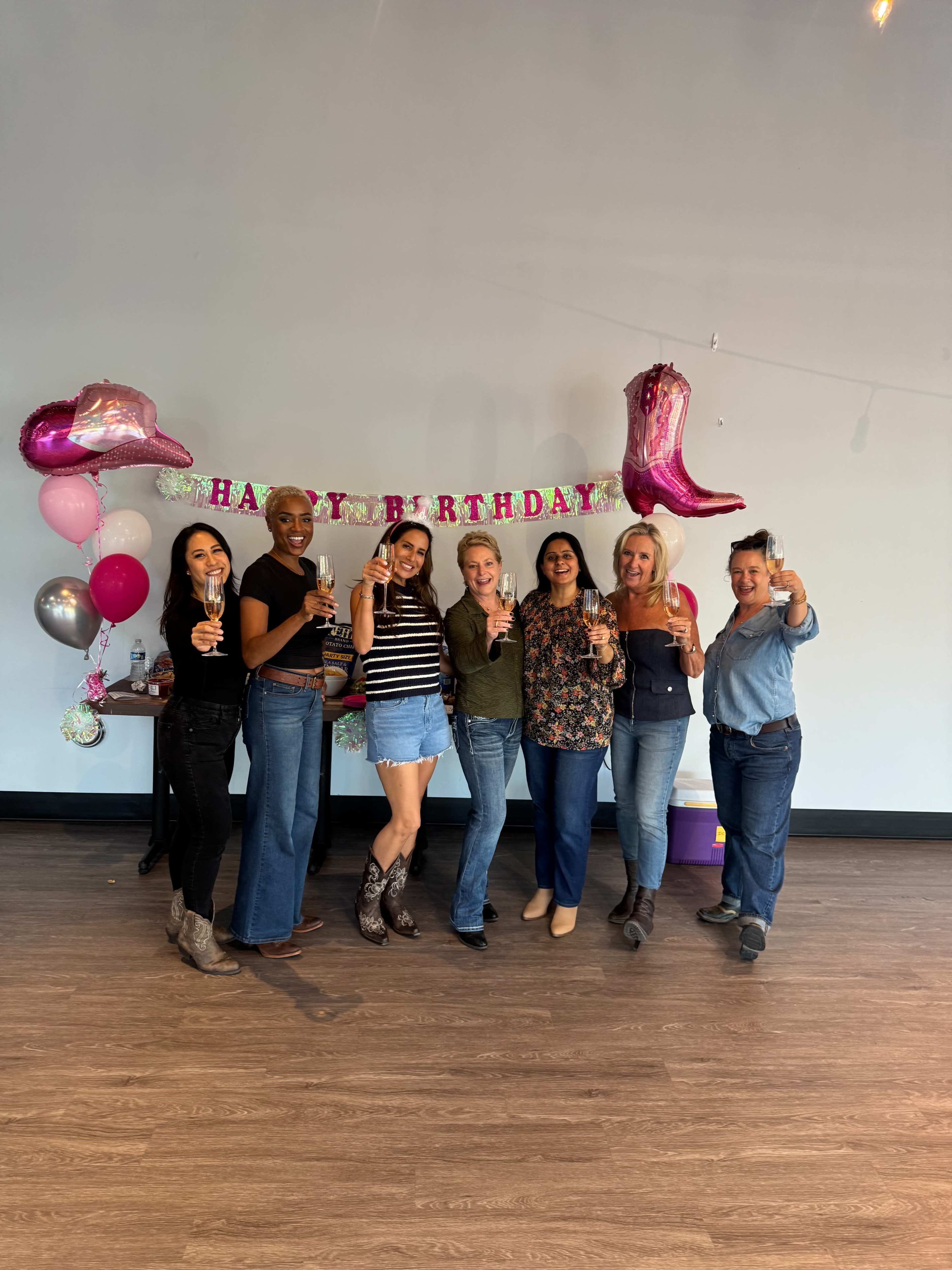 A group of seven women stands together, holding drinks in front of a "Happy Birthday" banner and decorated balloons.