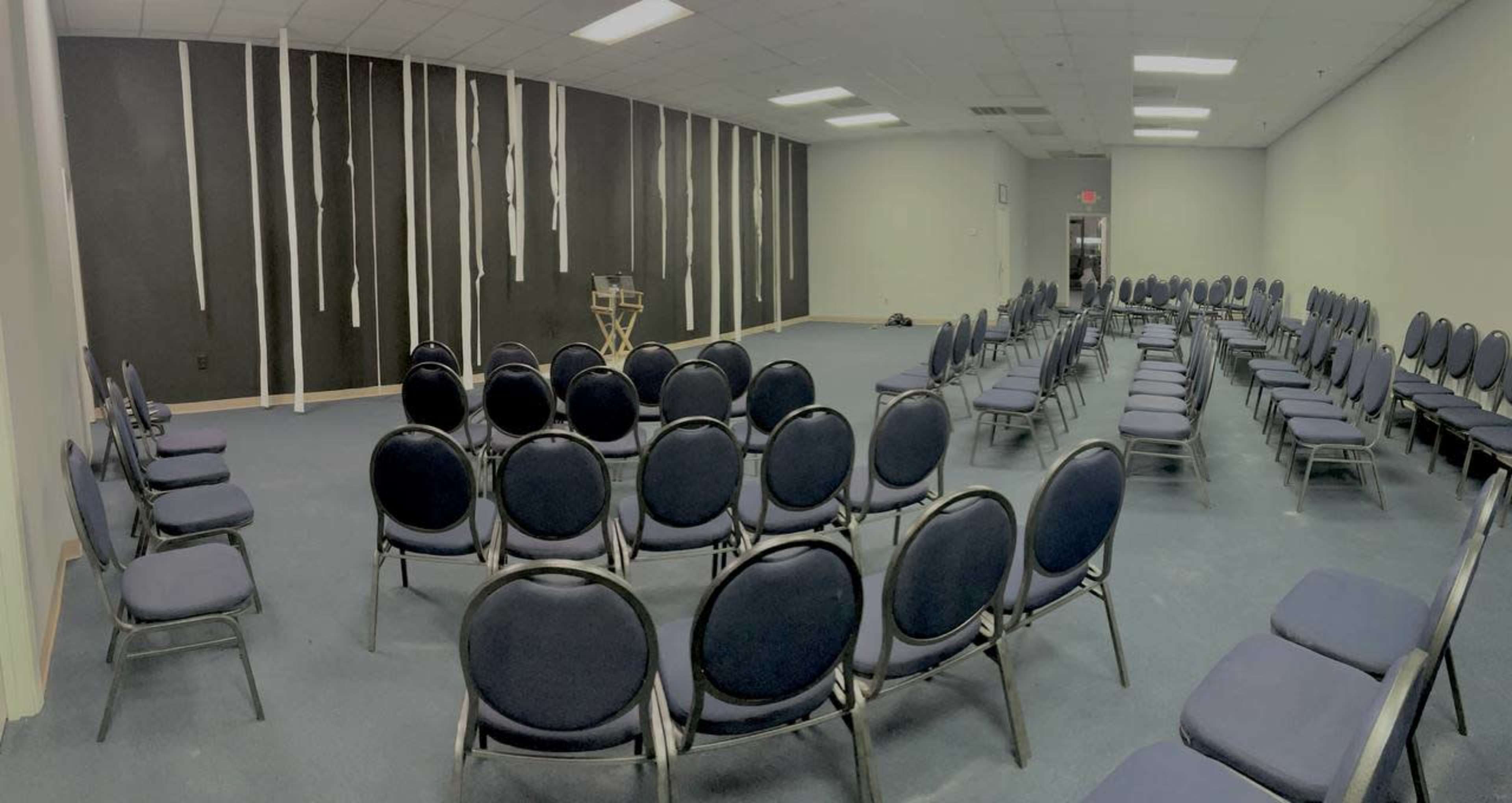The image shows a large, empty room with rows of gray chairs arranged in a circular pattern around a central space, with black walls and minimal decoration.