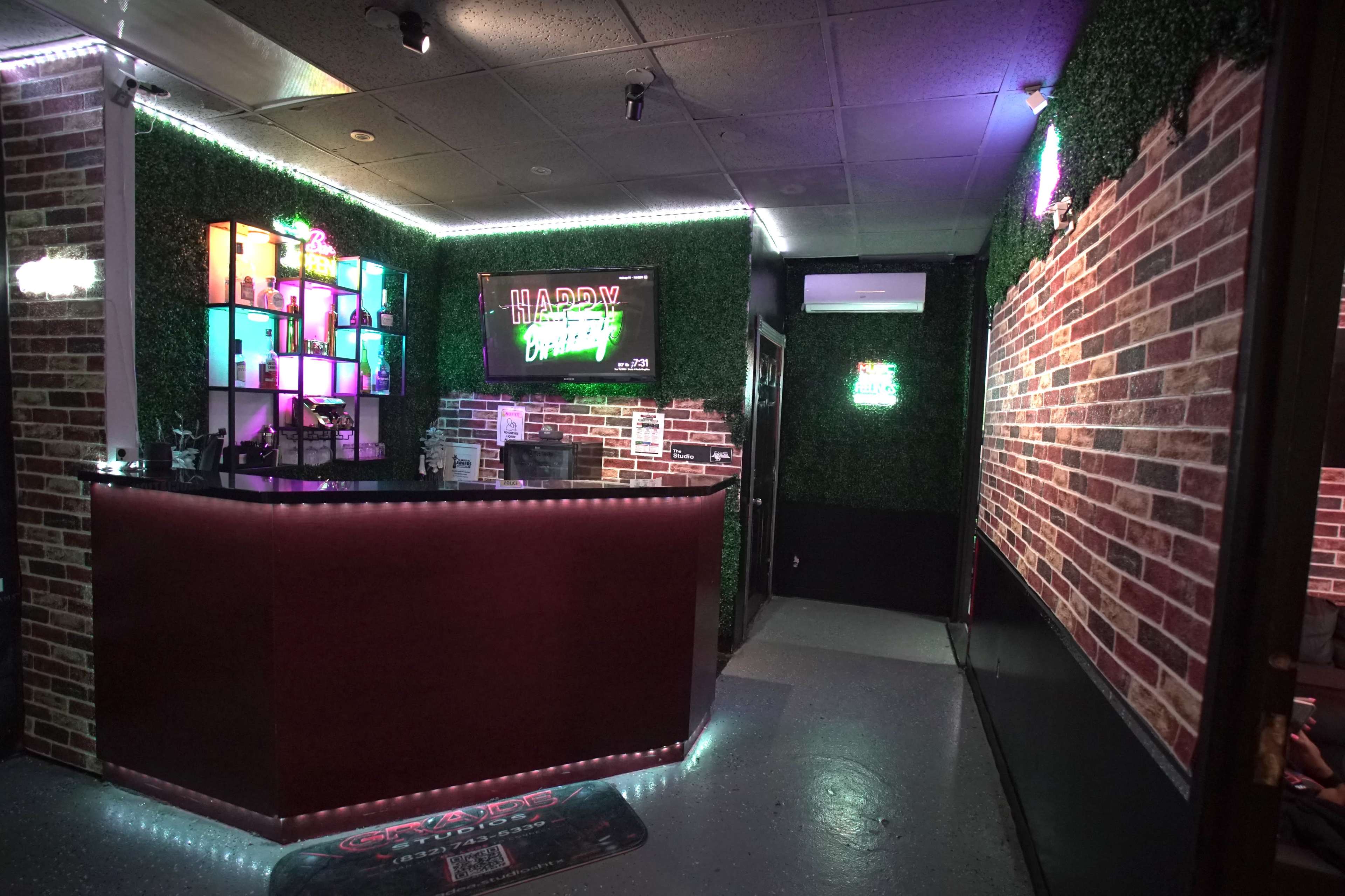 The image shows a brightly lit party venue with a reception area featuring a bar, colorful neon signs, and brick wall decor.