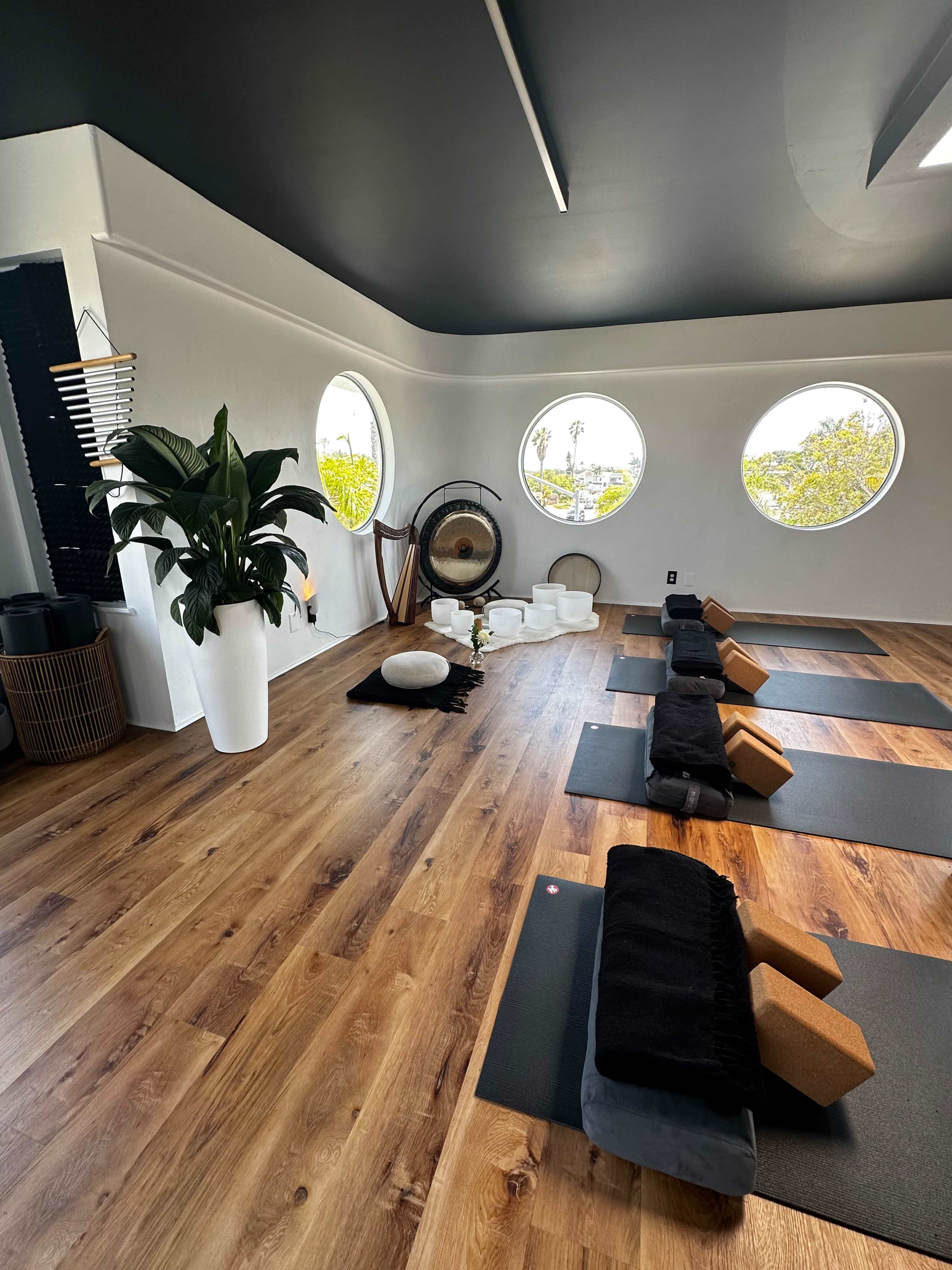 The image shows a serene yoga studio with circular windows, wooden flooring, and neatly arranged yoga mats and props.
