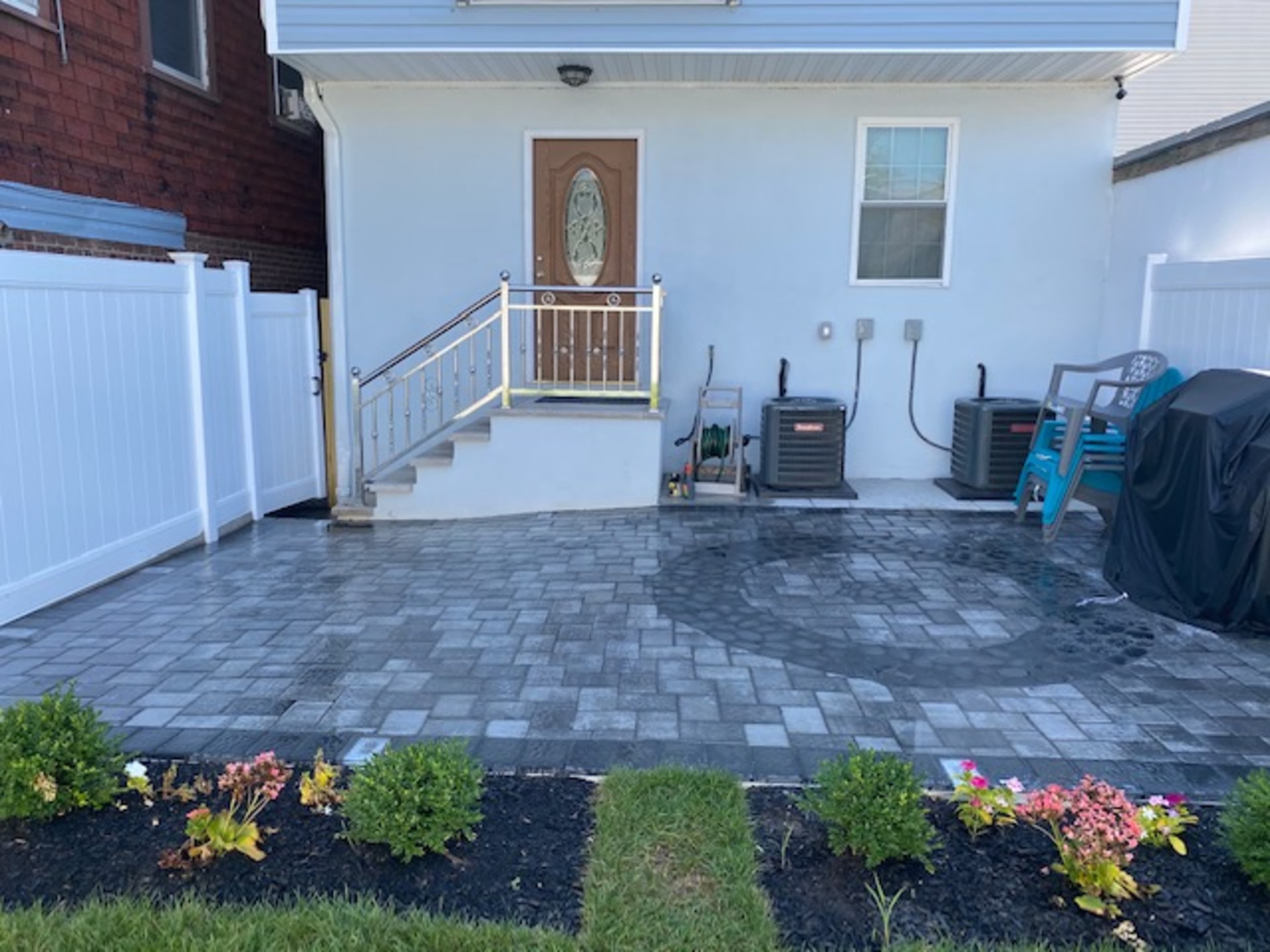 A paved patio area leading to the entrance of a house, with a staircase, air conditioning units, and a garden with neatly arranged shrubs and flowers.