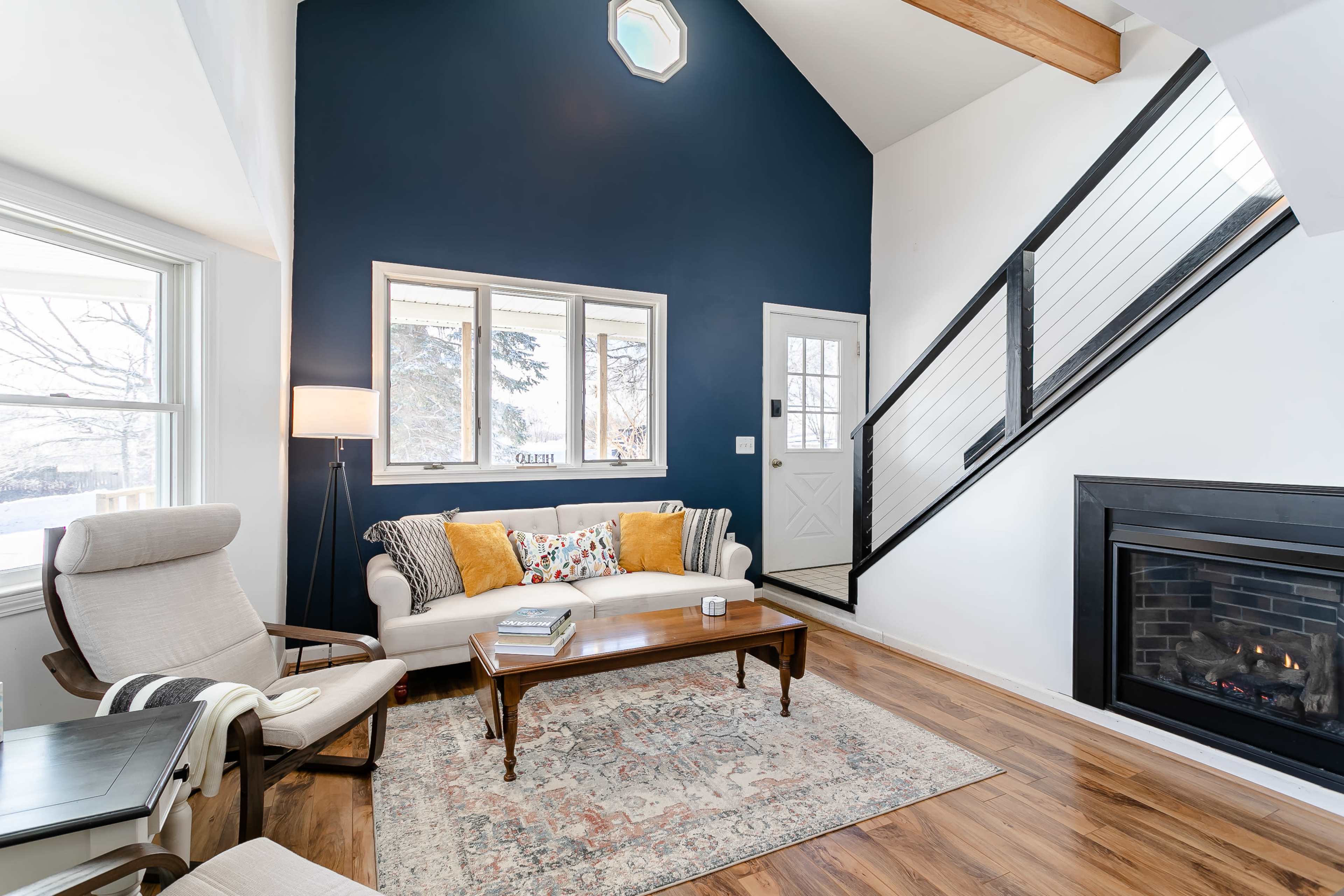 A modern living room with a white sofa, a wooden coffee table, and a fireplace, featuring large windows and a staircase in the background.