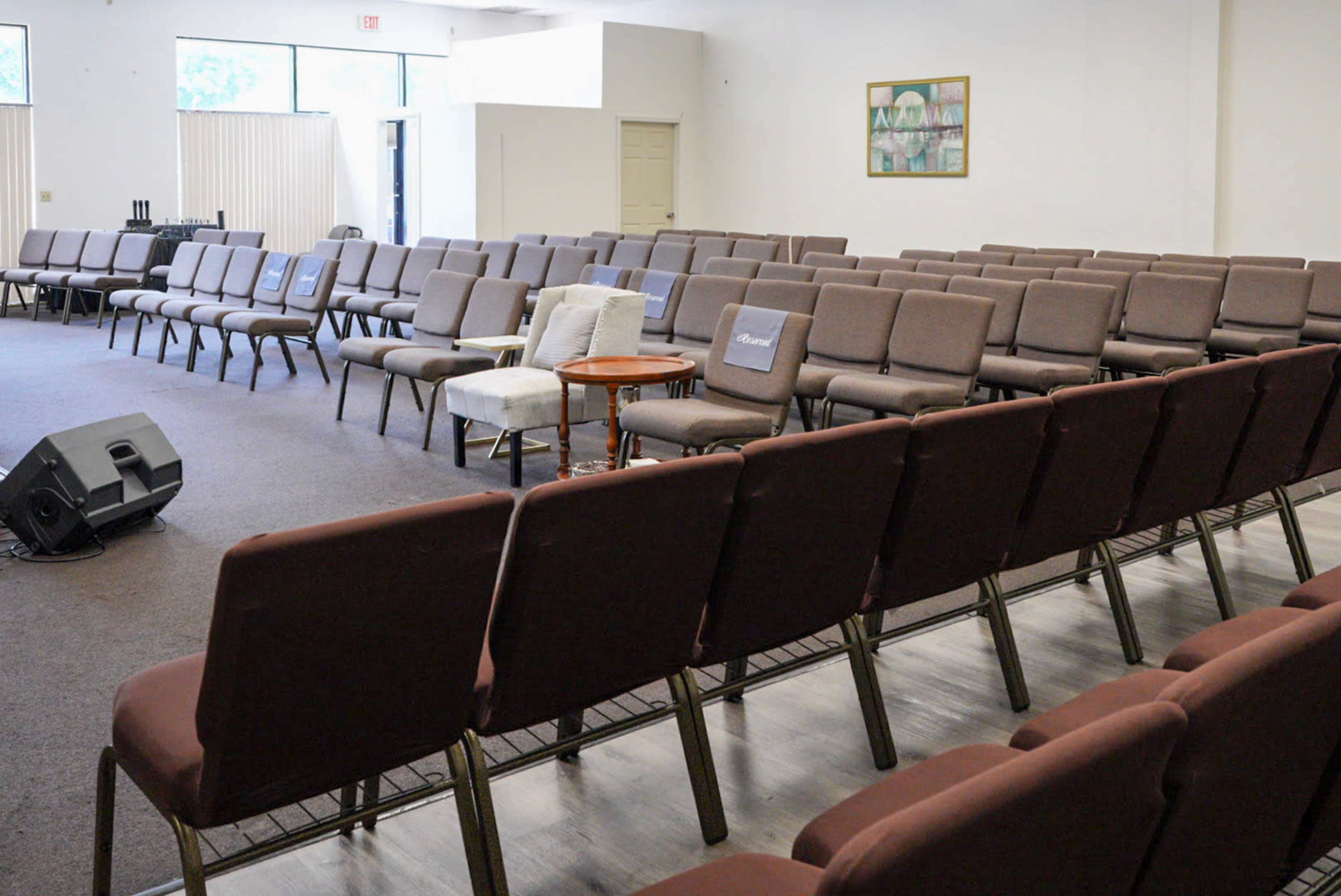 The image shows a row of empty, brown chairs arranged in a spacious room with light-colored walls and a small stage area.