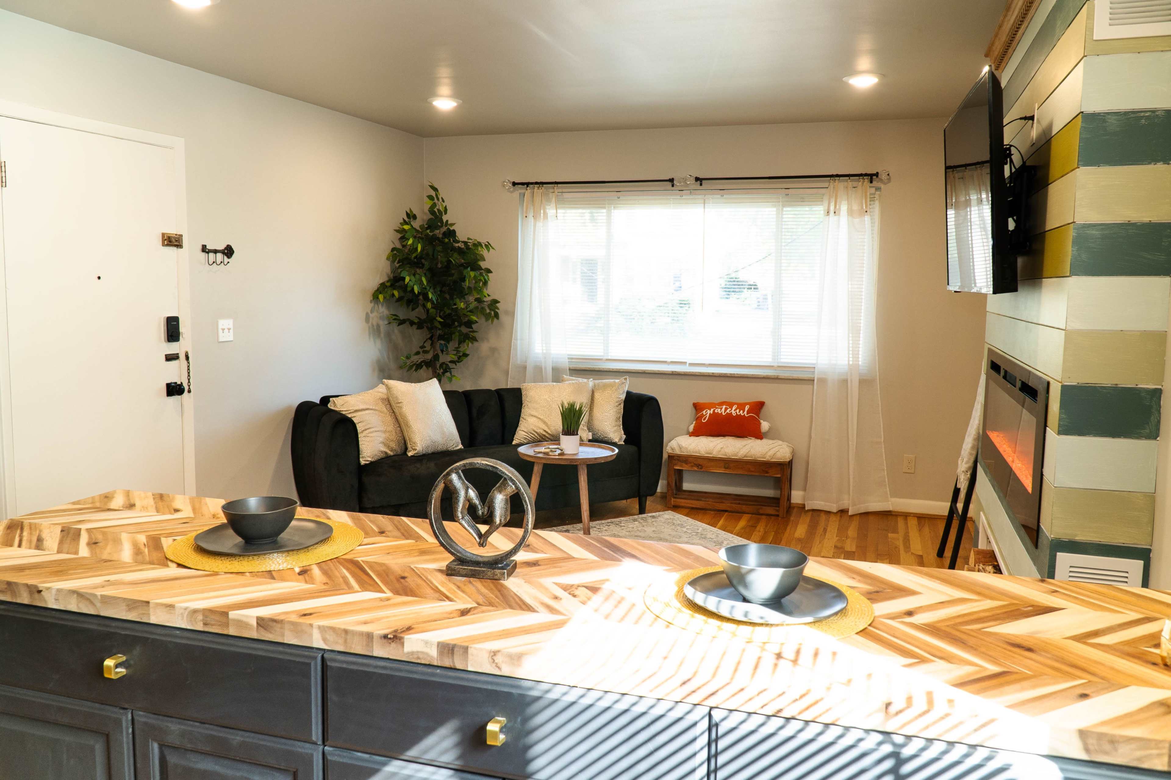 A modern living space features a dark green sofa, a small round table with a decorative plant, and a wall-mounted TV, all set against a backdrop of a well-lit window and a stylish countertop.