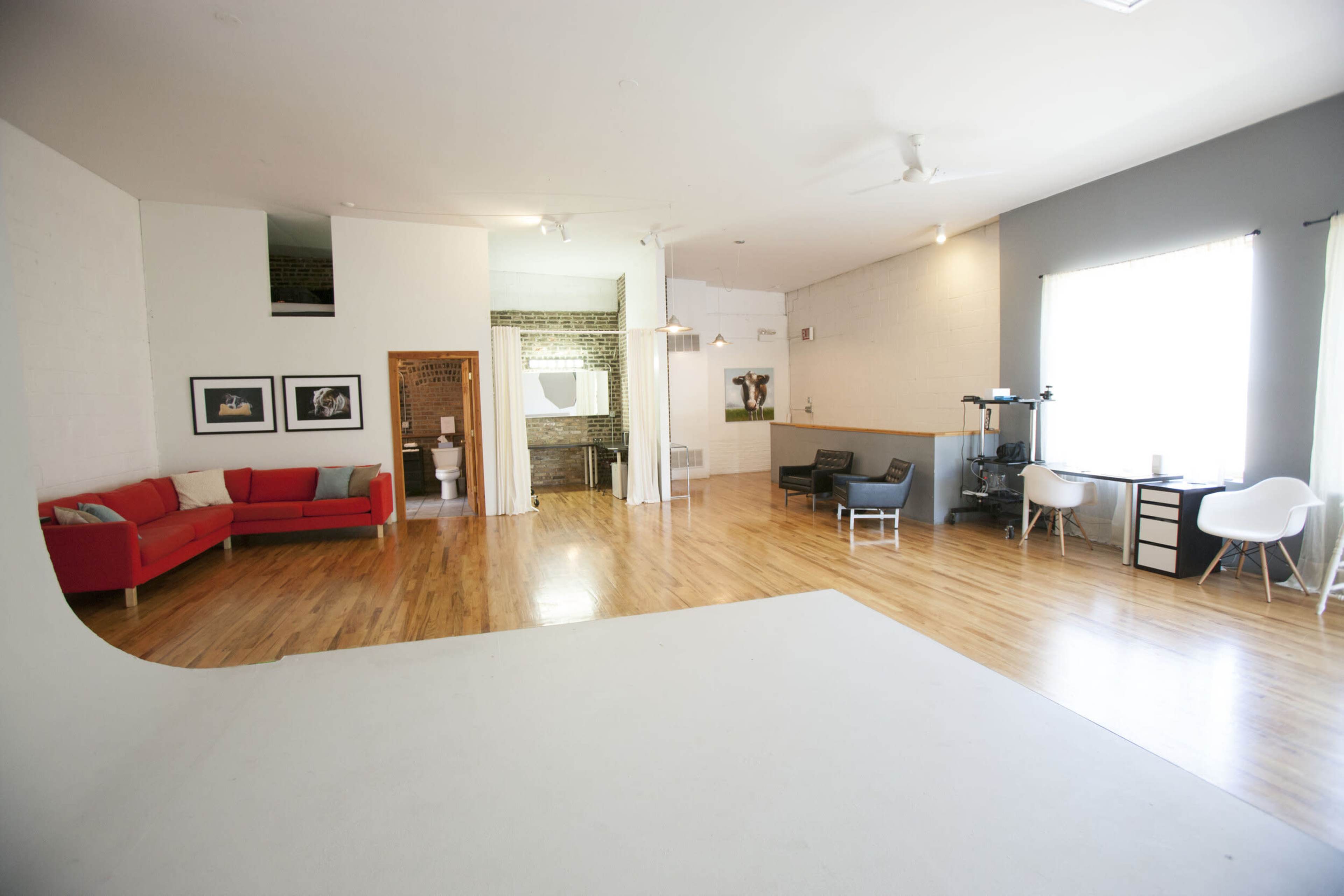 A spacious, minimalist studio with wooden flooring, a red couch, and separate areas including a bathroom and workspace.