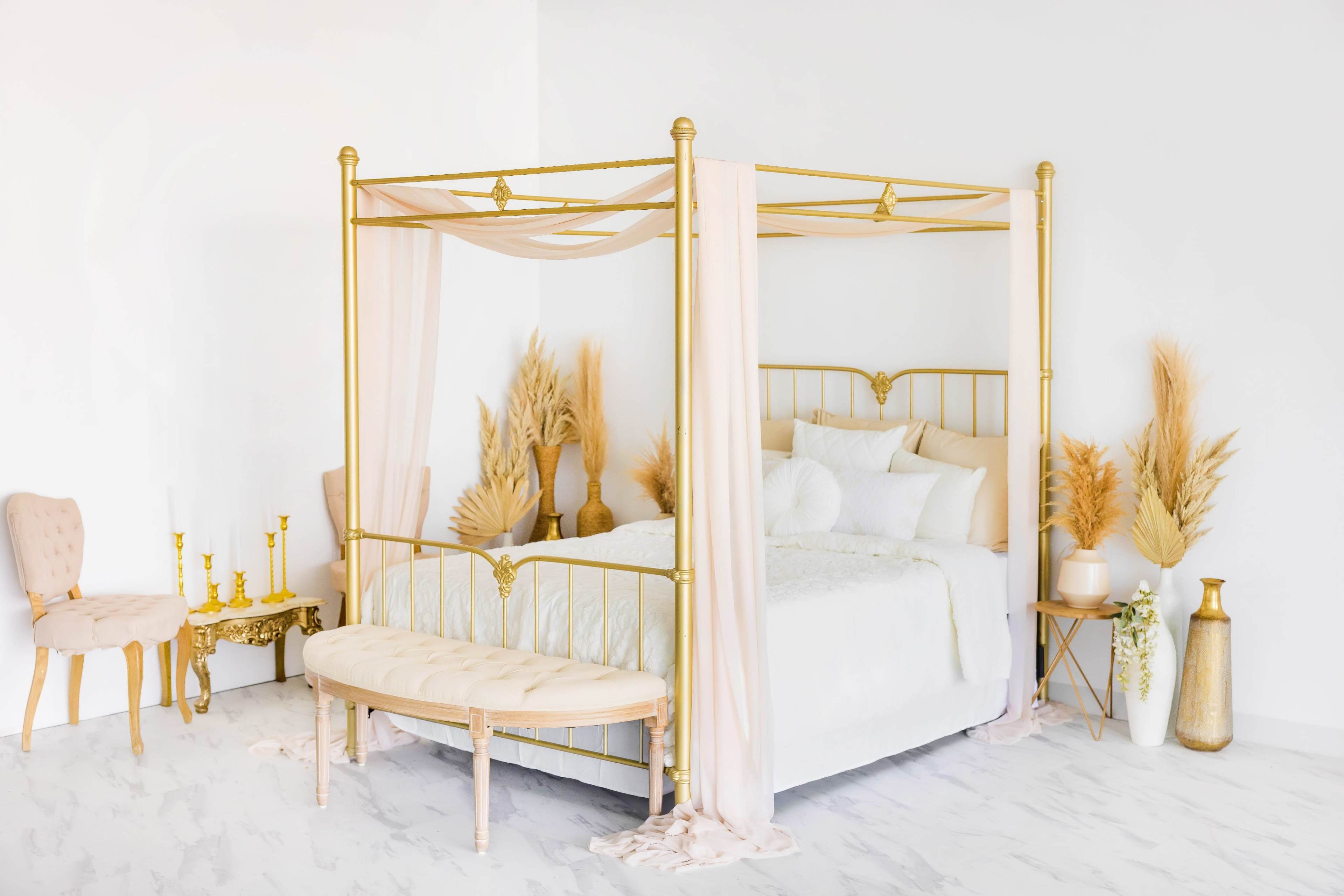 A gold metal four-poster bed with light fabric drapes, complemented by decorative plants and soft furnishings in a bright, minimalist bedroom setting.