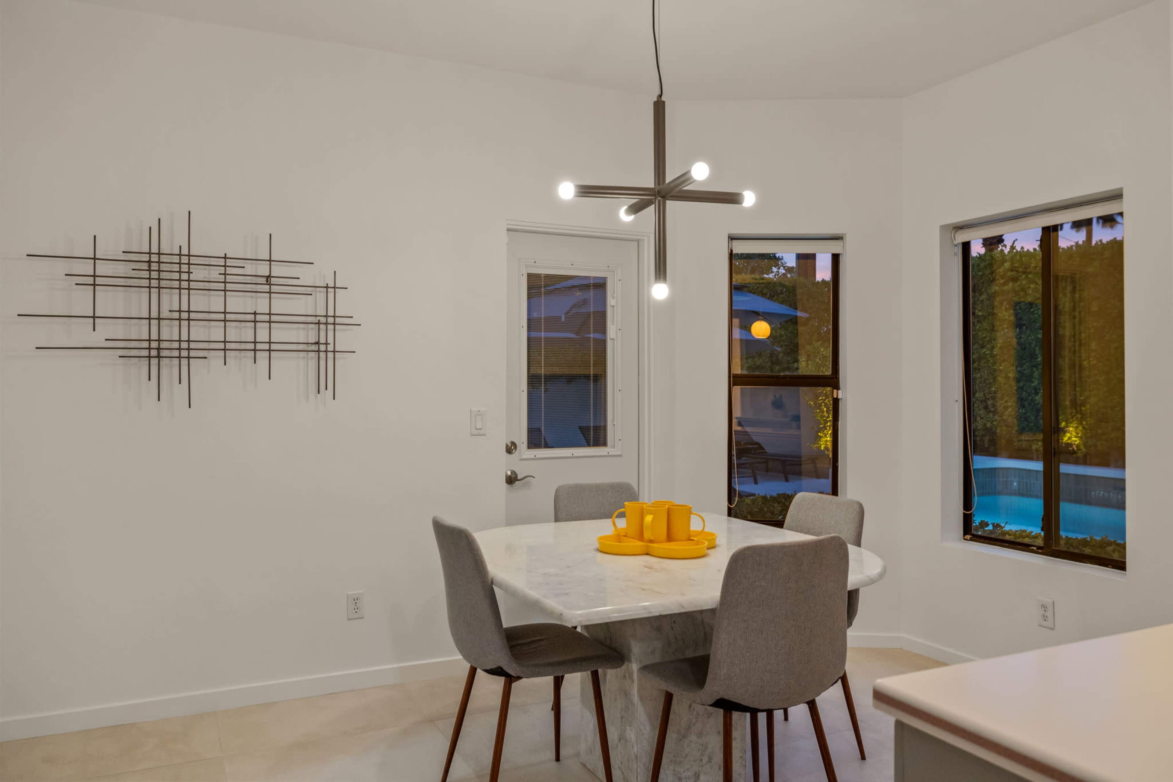 A modern dining area features a round marble table with grey chairs, illuminated by a geometric pendant light, and a view of a pool through large windows.