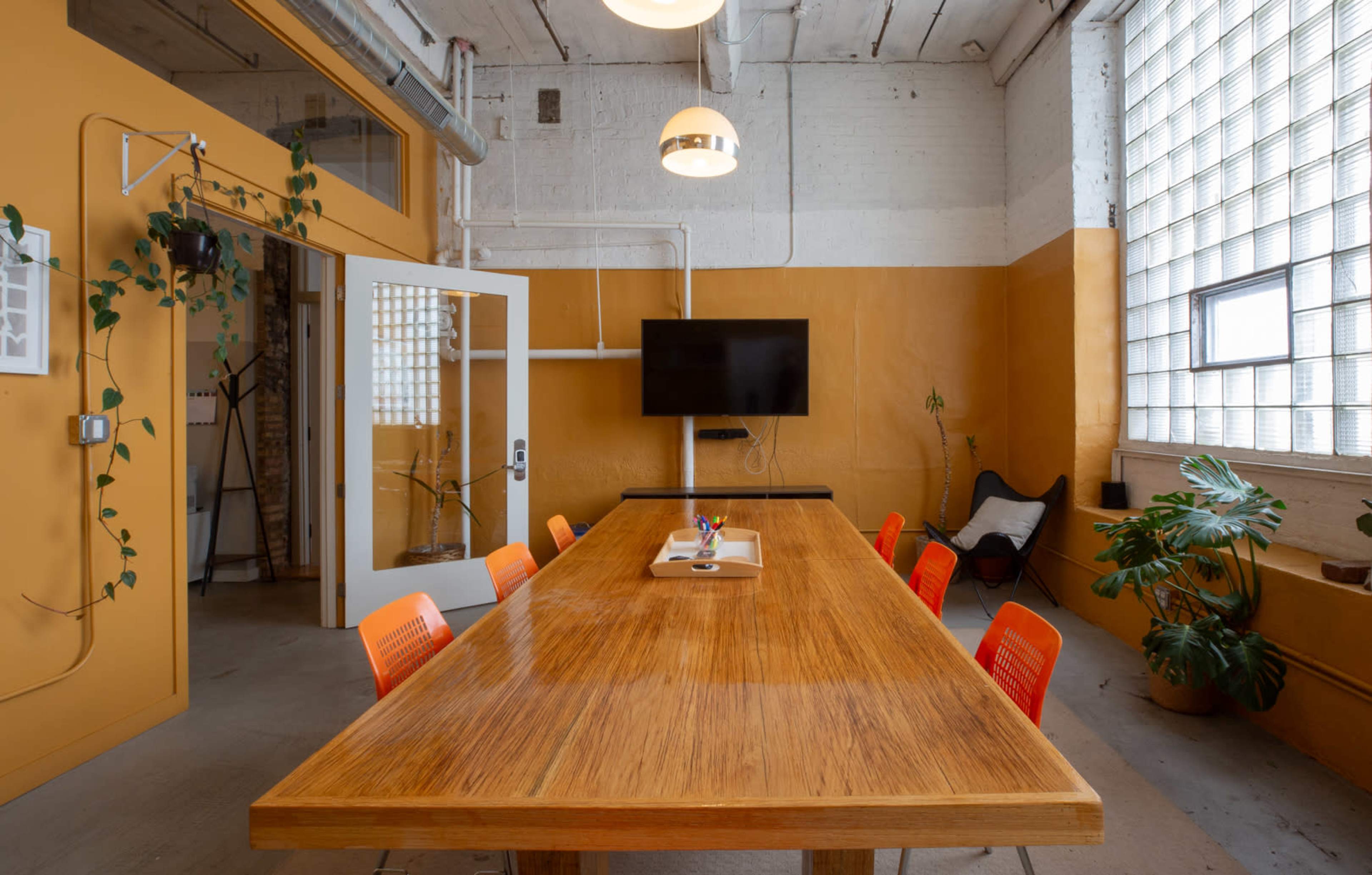 A large wooden conference table with orange chairs sits in a brightly colored meeting room featuring a TV and plants.