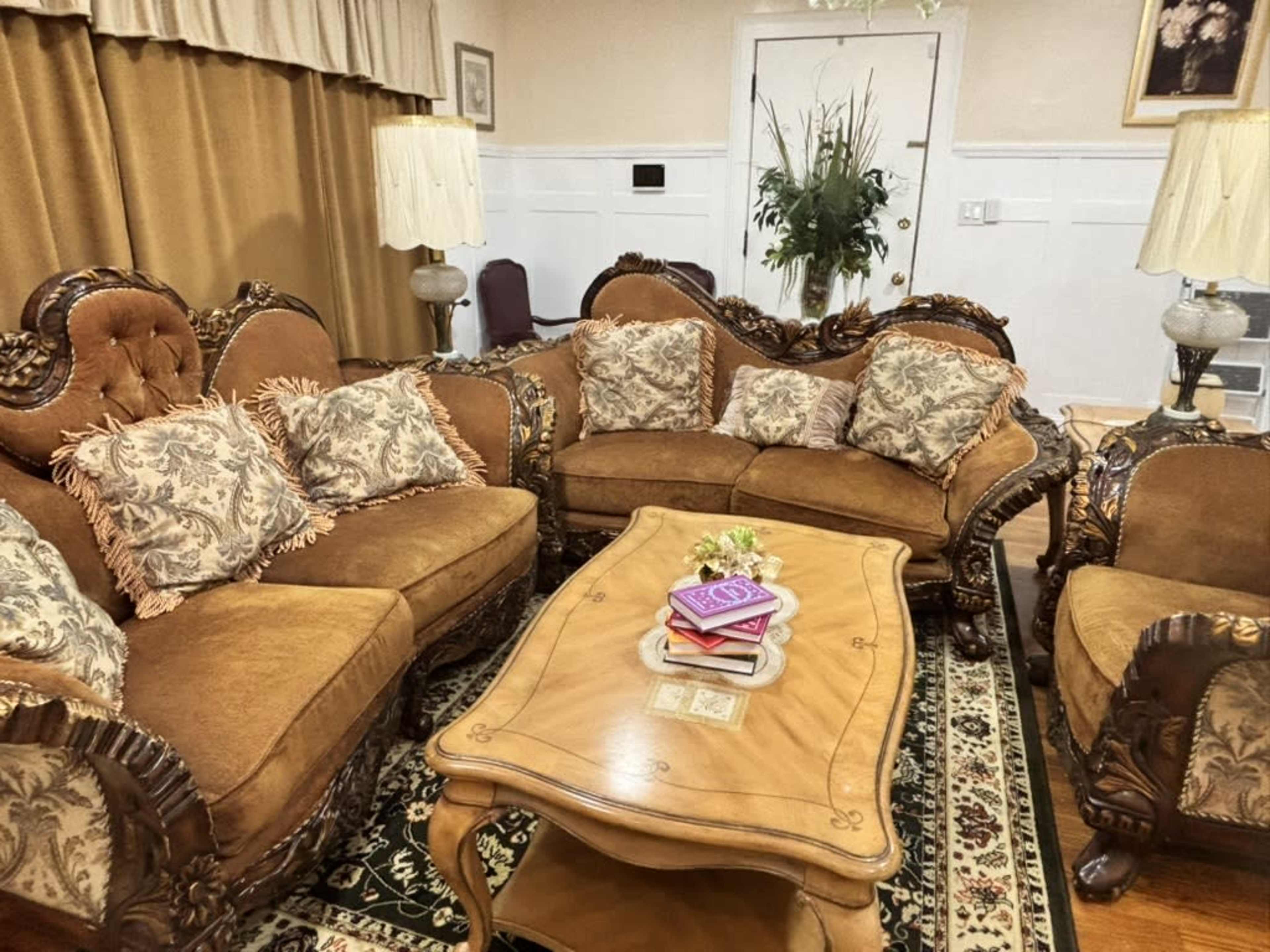 The image shows a furnished living room with a brown upholstered sofa set featuring ornate wooden detailing, a floral-patterned rug, and a coffee table adorned with books and a decorative flower arrangement.