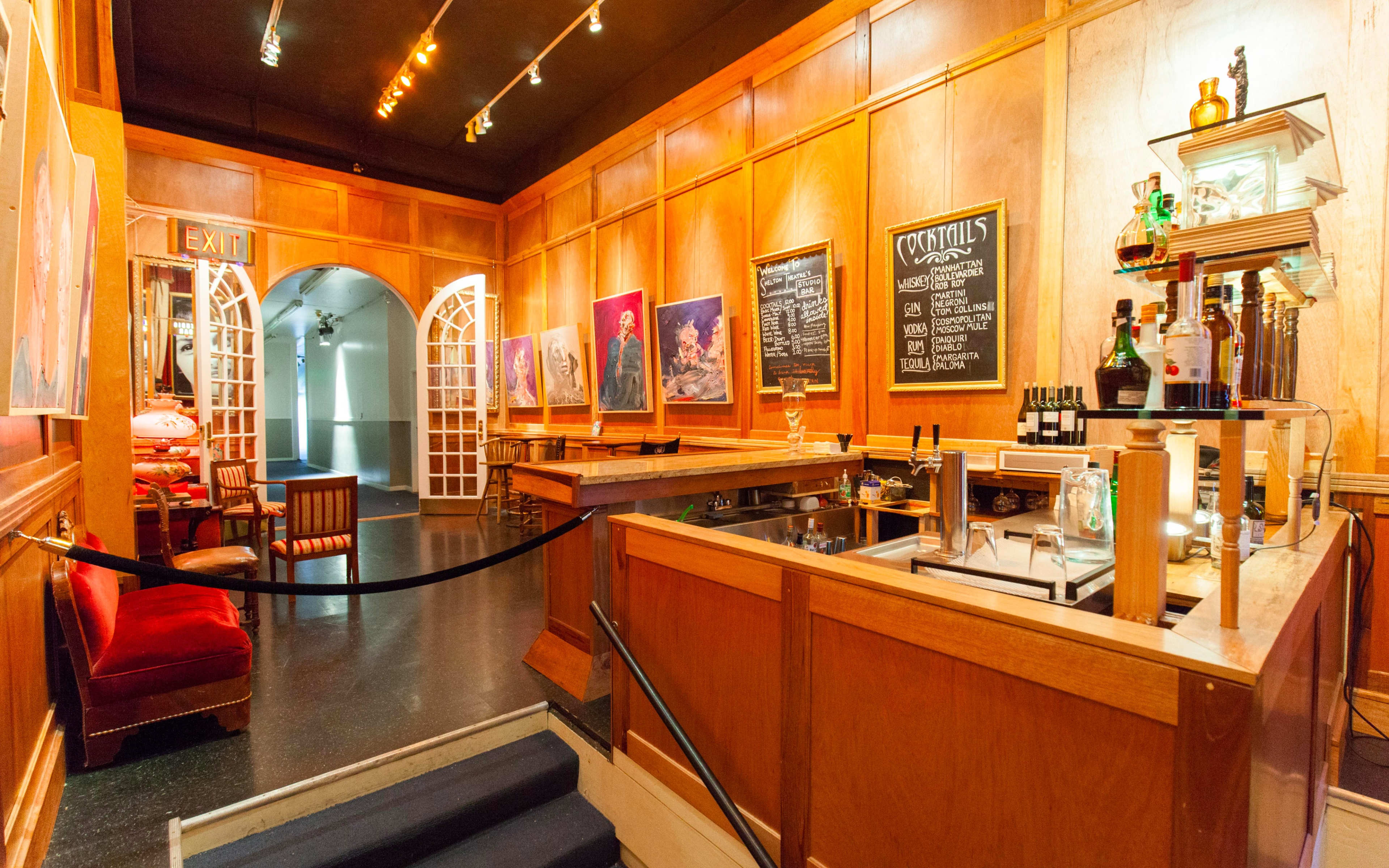 A wooden-paneled bar area with a counter, liquor bottles, and a chalkboard menu, leading to an arched exit adorned with a few framed artworks.