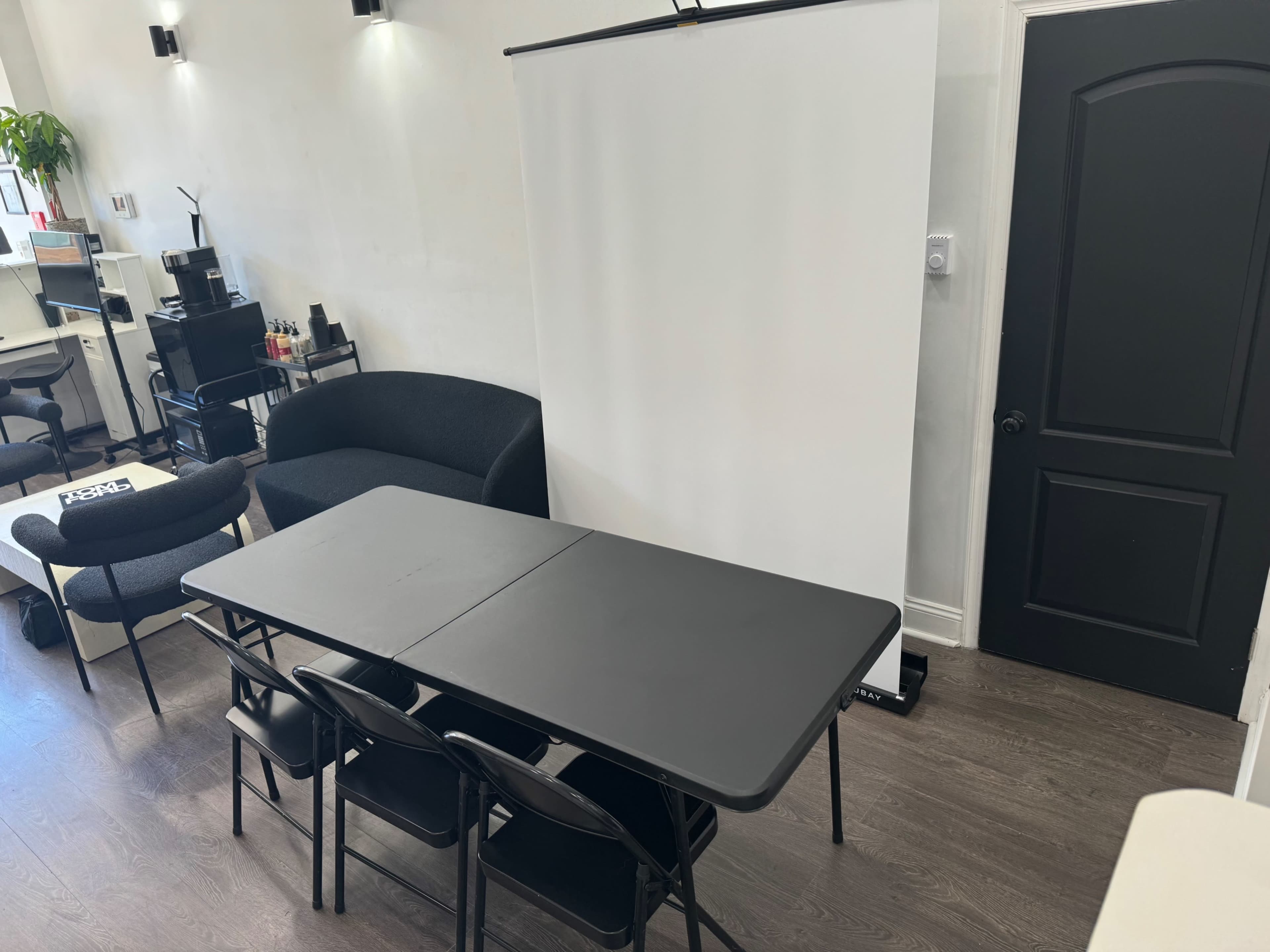A room with a black folding table and four chairs arranged in front of a white projection screen, with a dark door visible at the side.
