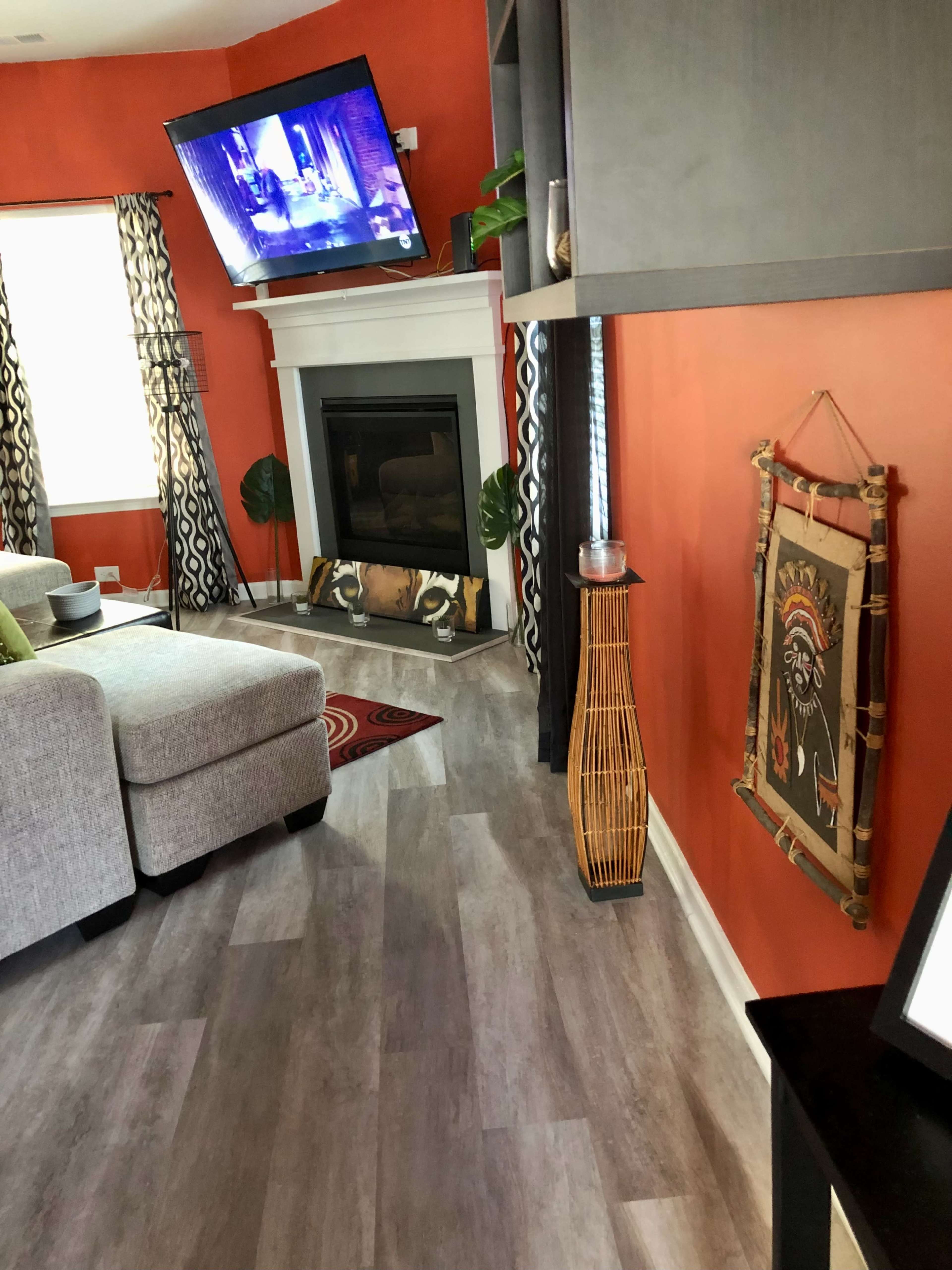 A modern living room features a gray sectional sofa, a flat-screen TV mounted on a bold orange wall, and decorative accents including a tall woven light fixture and a wall hanging.