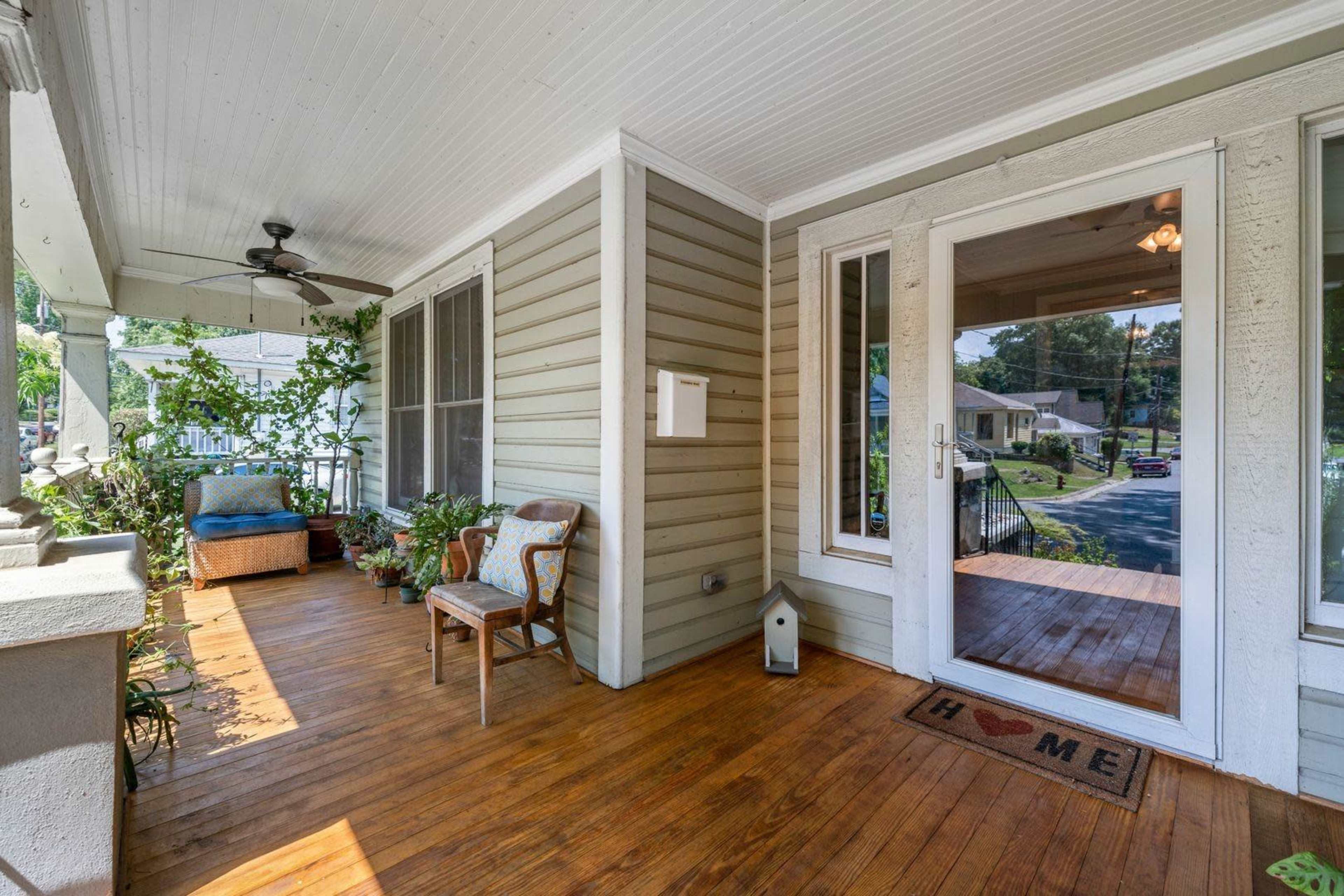 A front porch with wooden flooring, a chair, and potted plants, leading to a door with a glass panel.