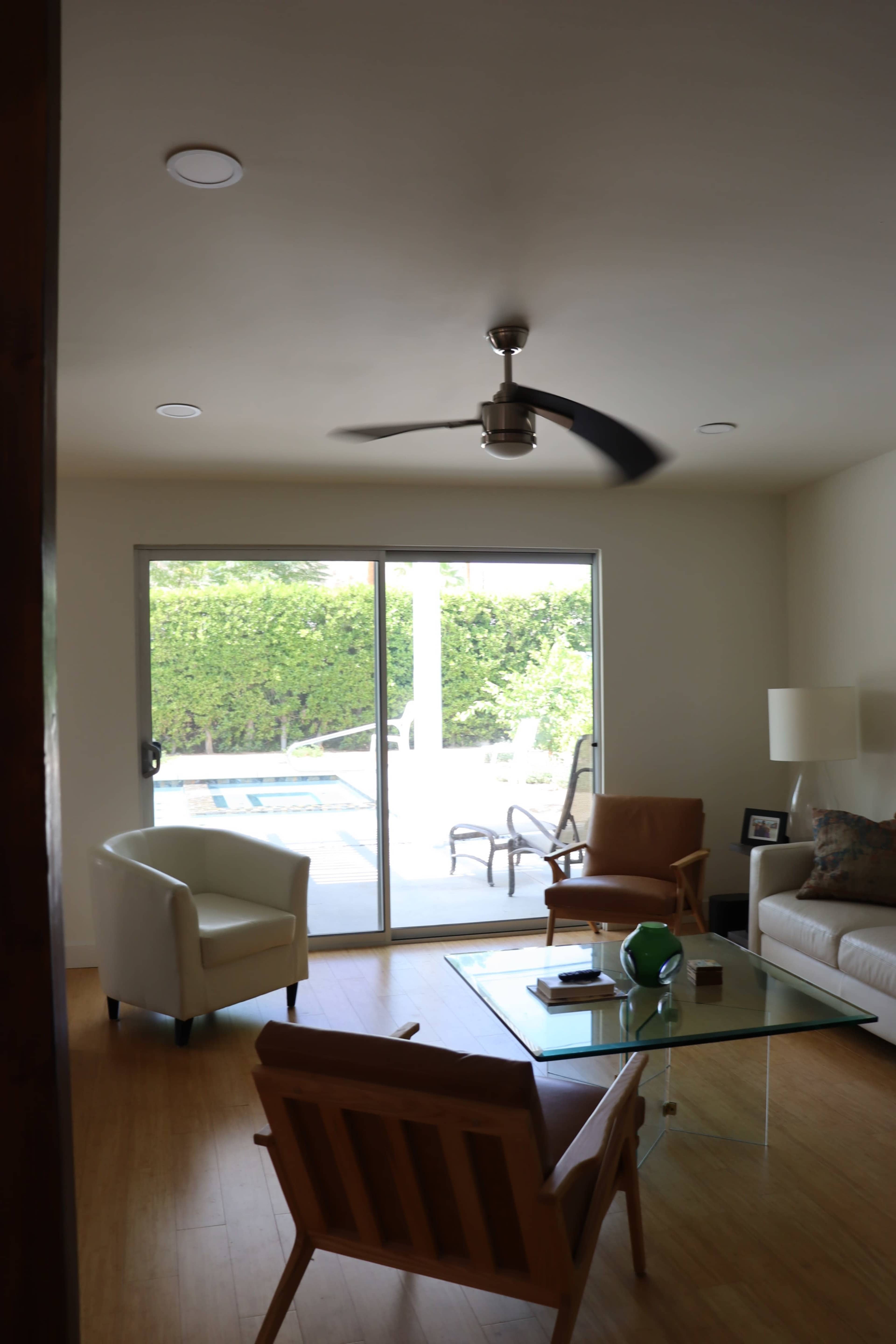 The image shows a living room with a glass coffee table, a white armchair, an orange-brown chair, and a large sliding door leading to a pool area.