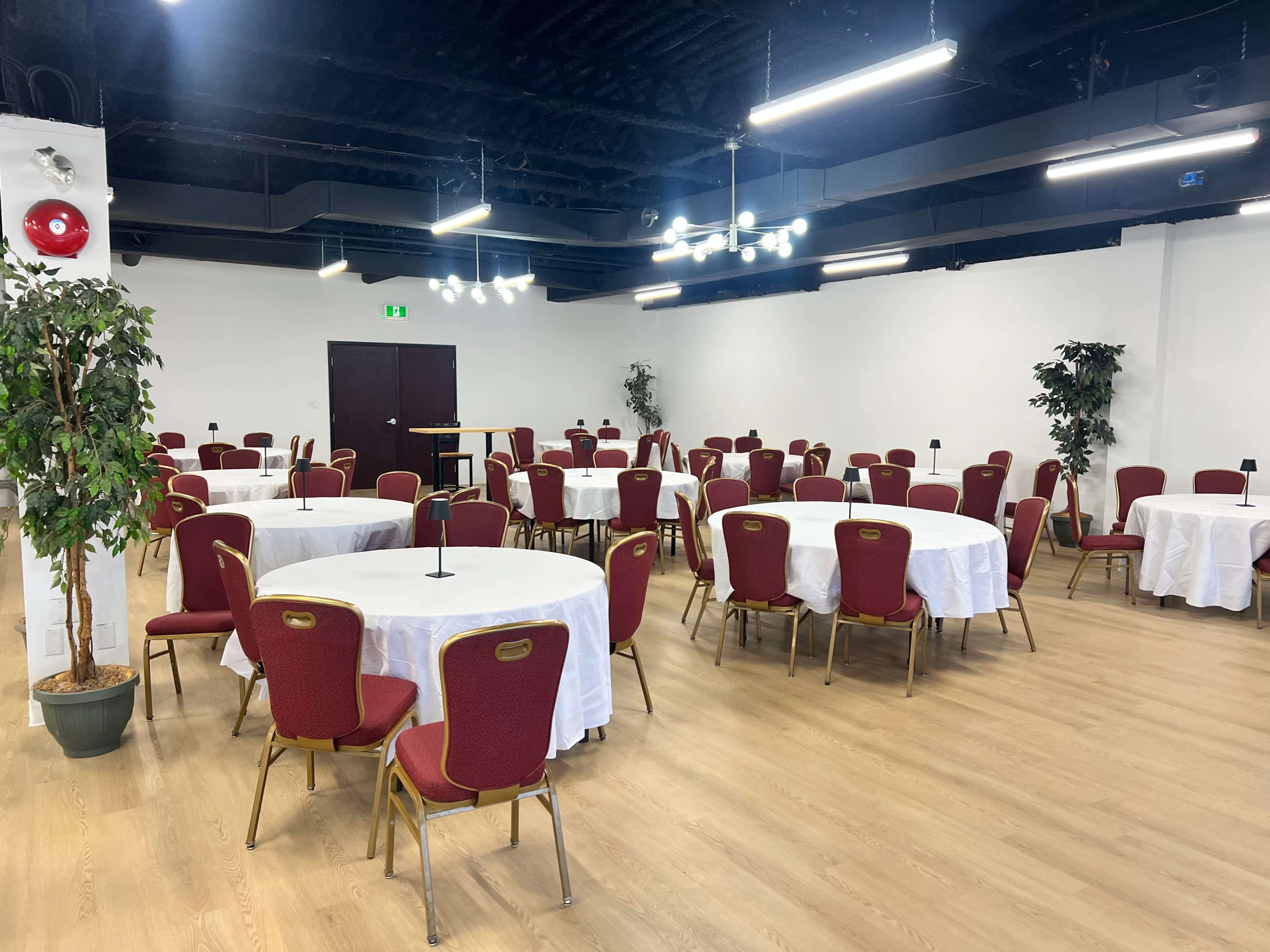 The image shows a spacious event room with several round tables covered in white tablecloths and red chairs arranged neatly throughout the area.
