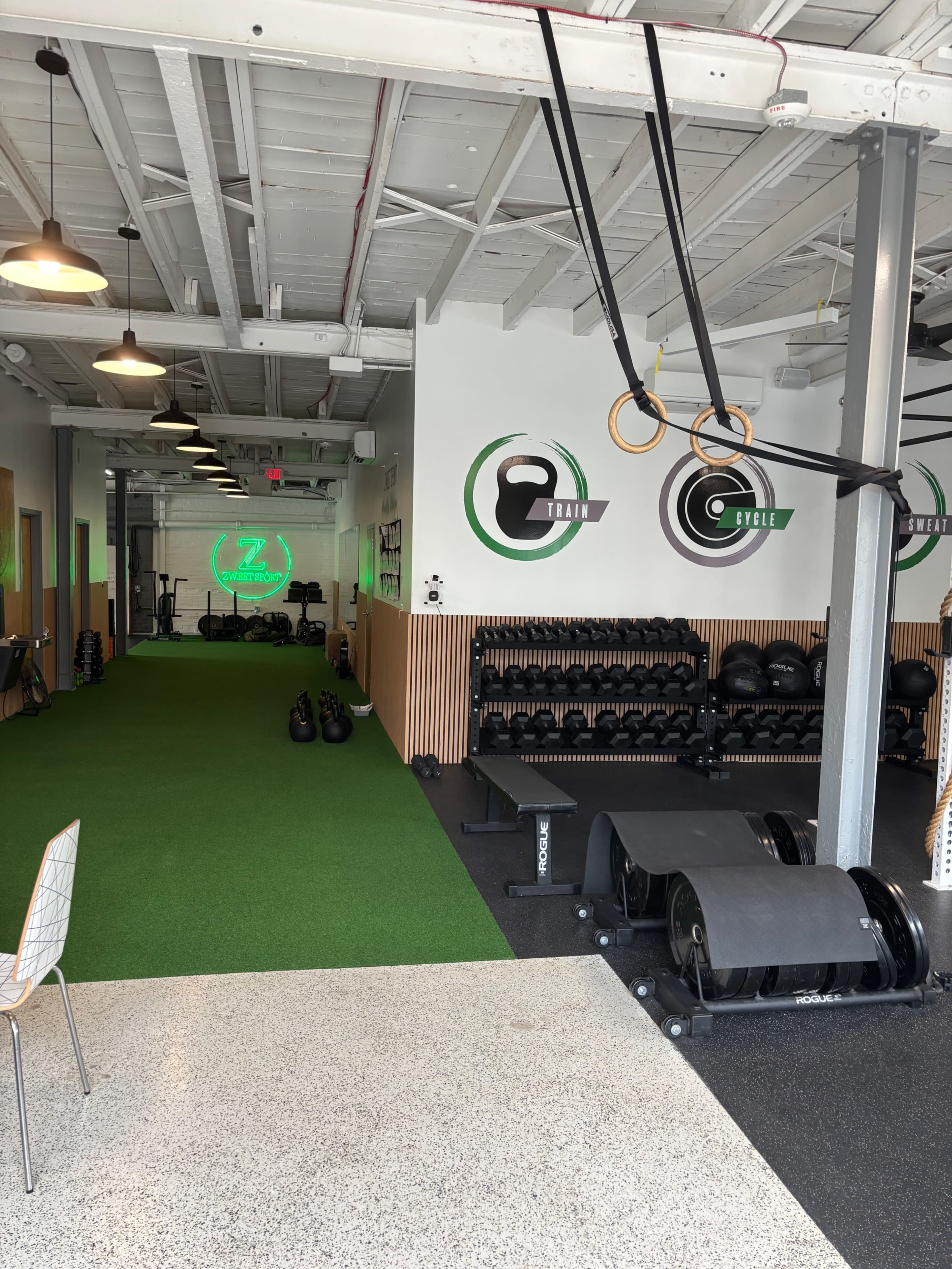 The image shows a modern gym interior with green turf flooring, weightlifting equipment, and wall-mounted rings.