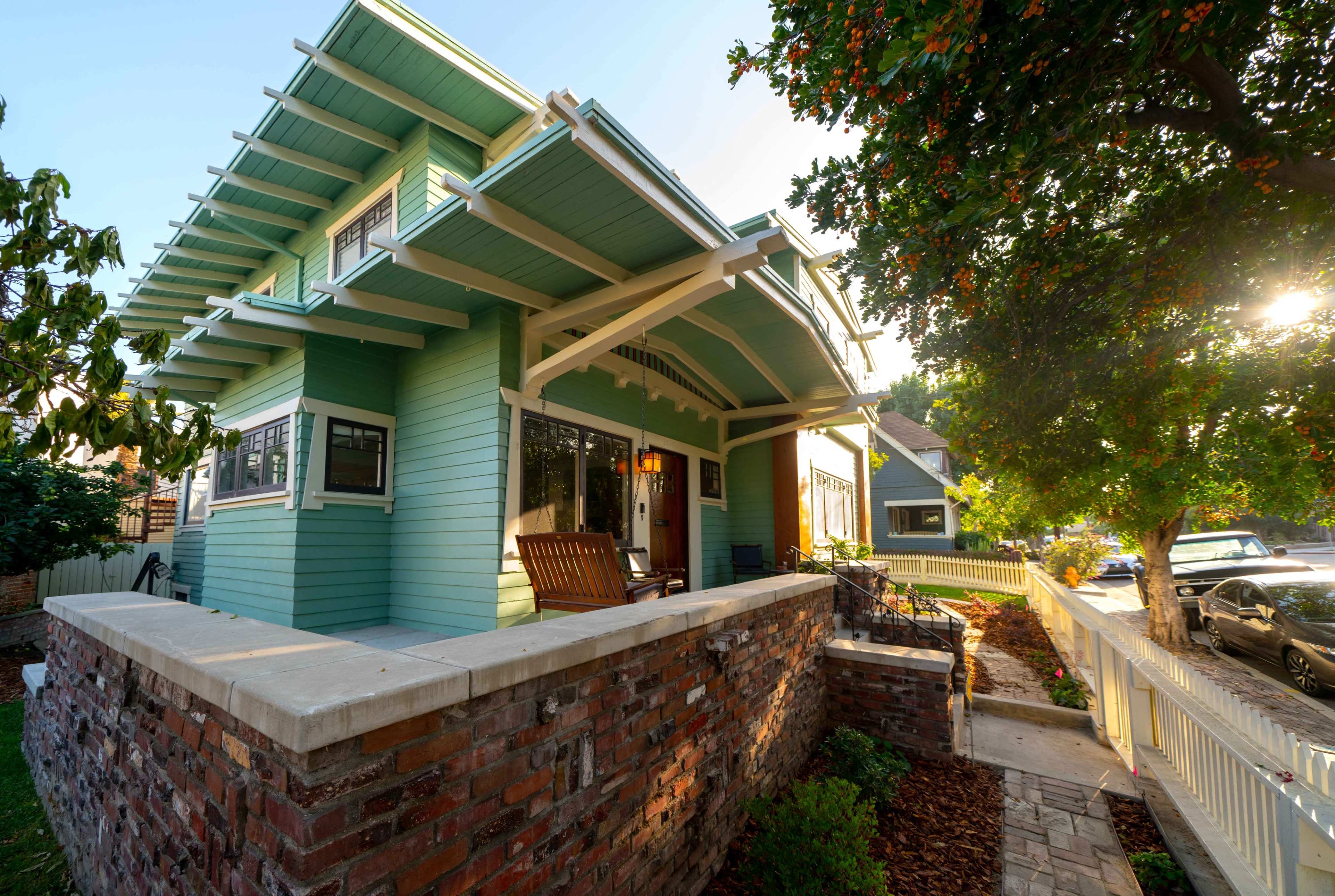 A blue-green Craftsman-style house with a front porch and seating area is situated on a corner lot, surrounded by landscaped gardens and parked cars.