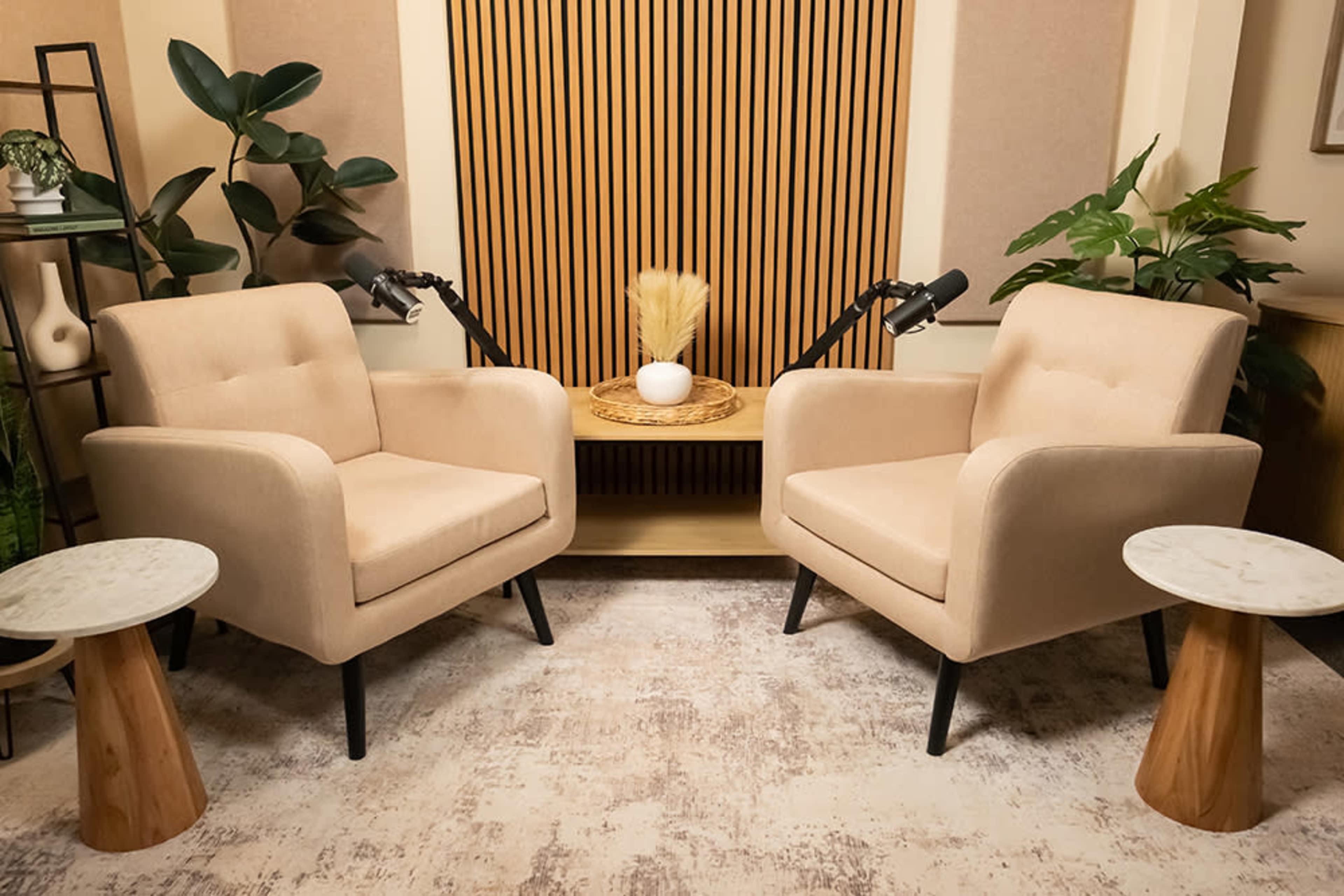 The image features two beige armchairs facing each other, placed on a textured rug, with two small round tables beside them and decorative plants in the background.