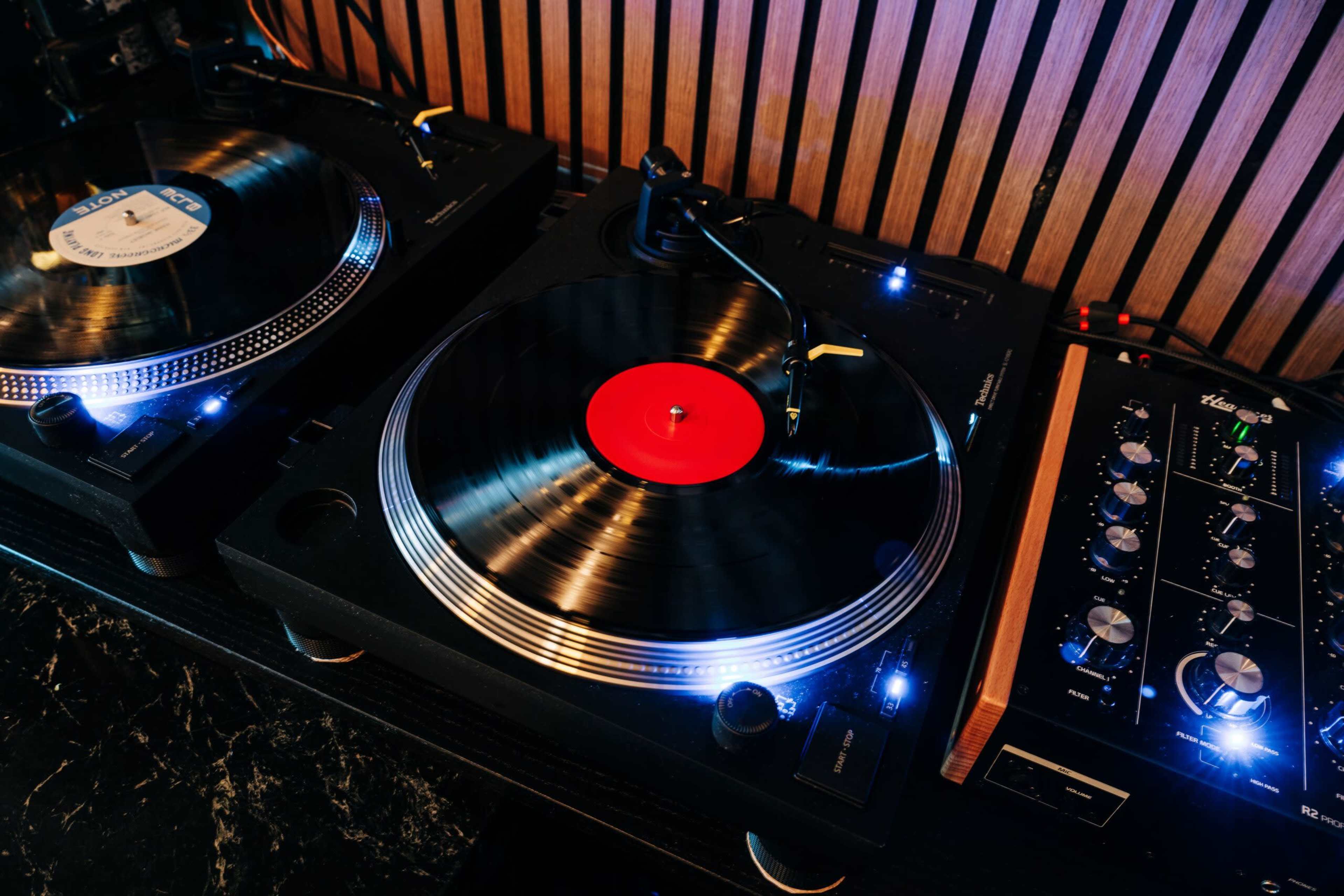 The image shows two turntables with one featuring a black vinyl record with a red label, positioned next to a mixer with knobs and lights.