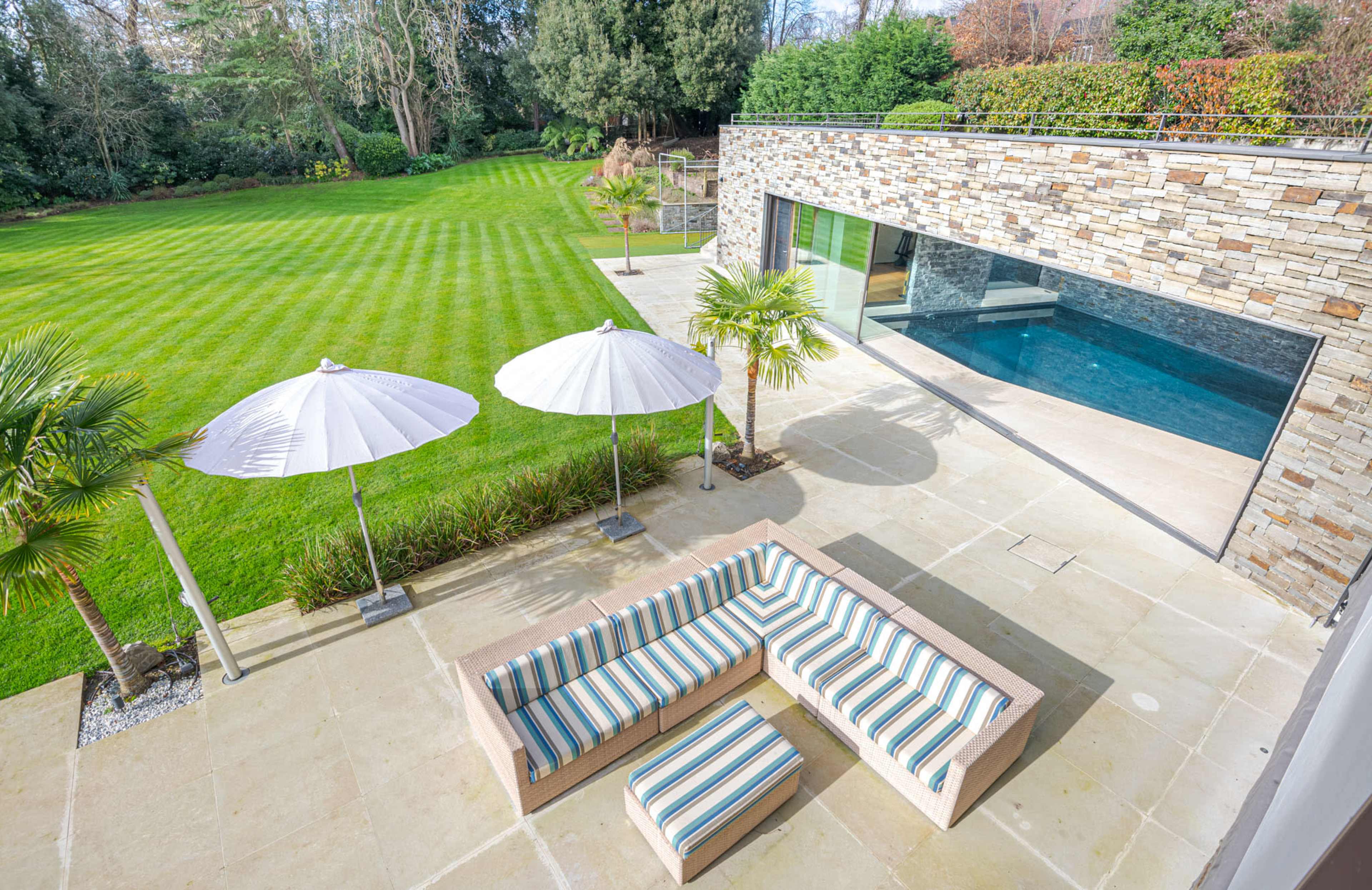 A modern outdoor patio area with striped seating, white umbrellas, and a pool adjacent to a landscaped lawn.