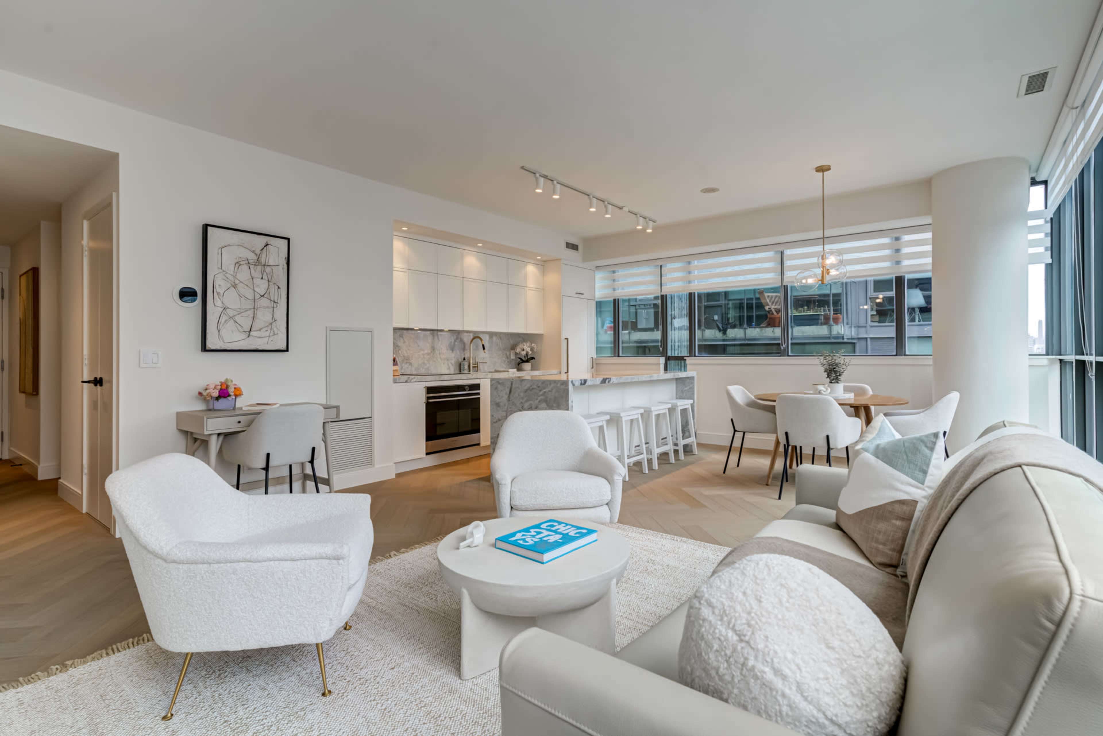 A modern open-concept living room and dining area with a light color palette, featuring a sofa, armchairs, a dining table, and a kitchen with contemporary cabinets.