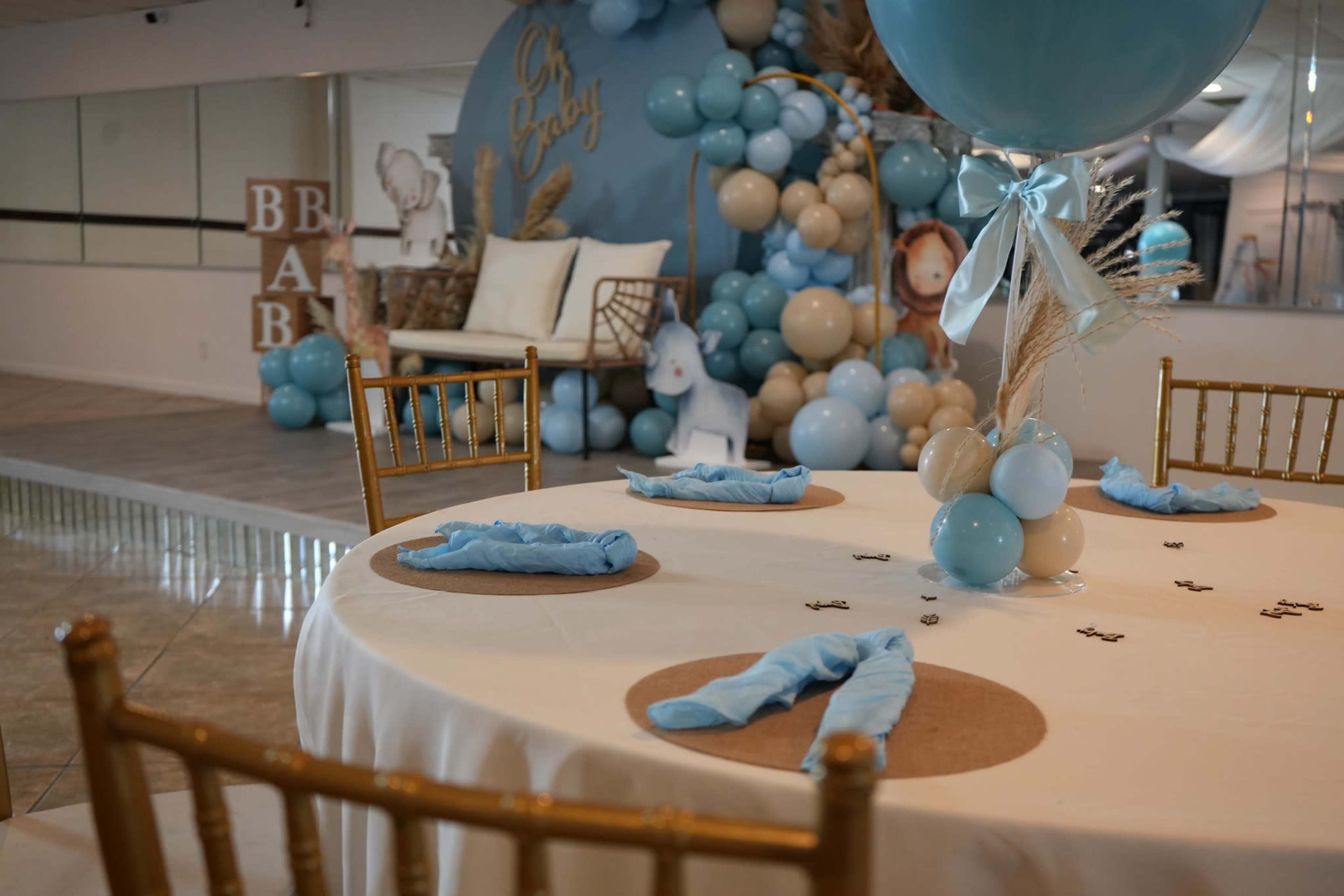 The image shows a decorated event space for a baby shower, featuring a table set with light blue napkins and a backdrop of balloons and a sofa.