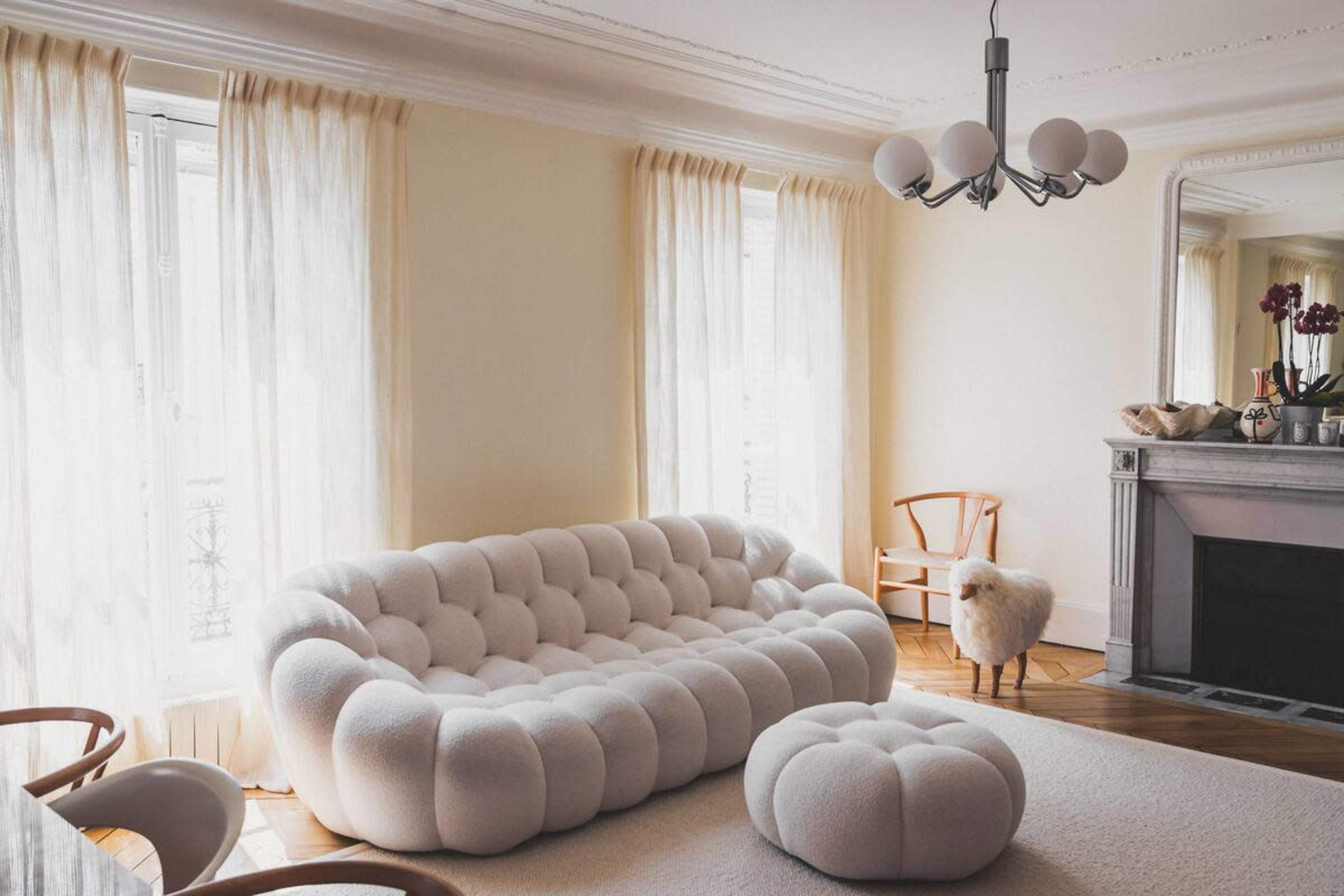 A living room featuring a white, tufted sofa, a matching ottoman, a wooden chair, and a fireplace with a decorative mantel.