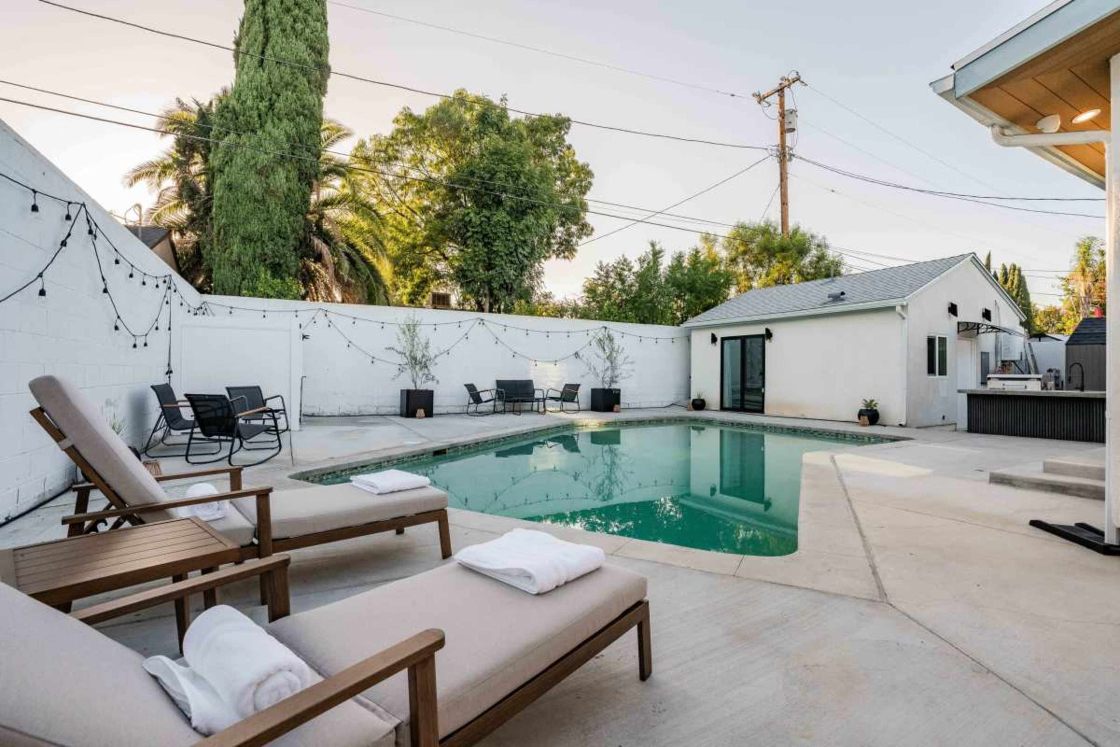 The image shows a backyard pool area with lounge chairs and a small building in the background surrounded by greenery.