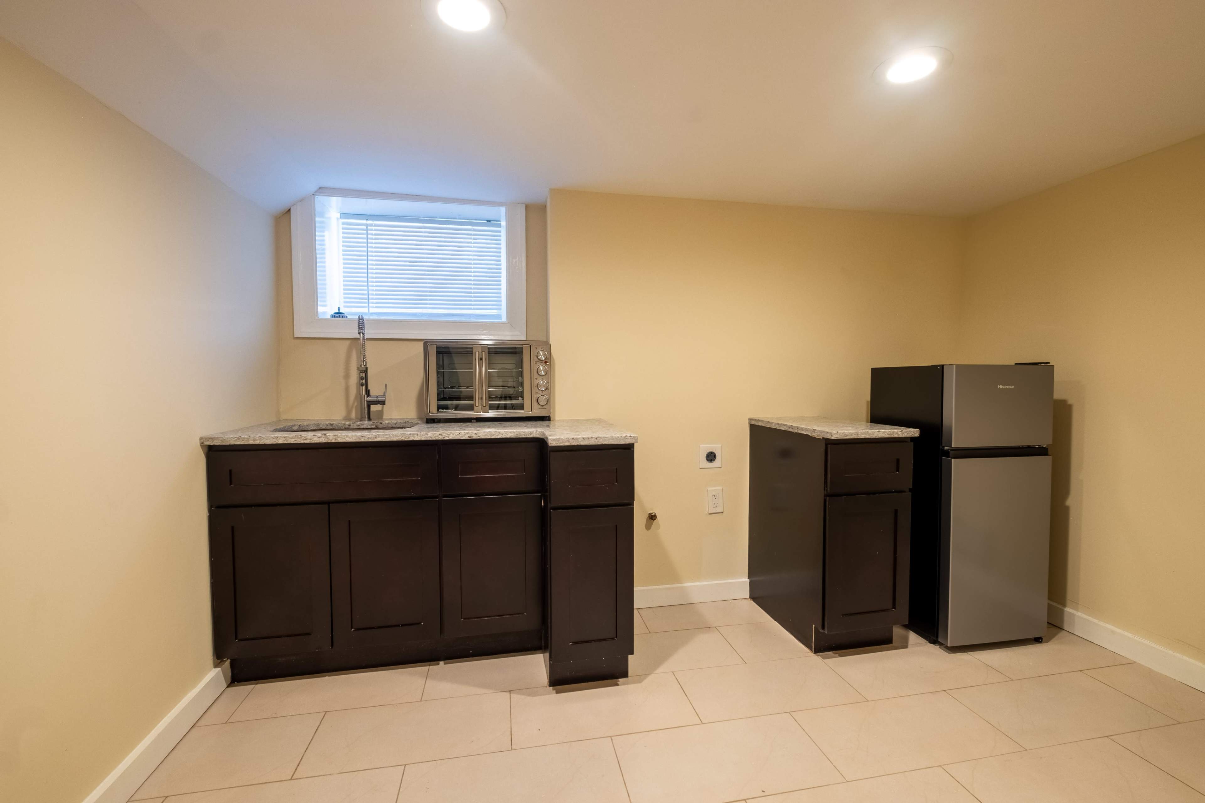A small kitchen area with dark wood cabinets, a countertop, a sink, a microwave, and a stainless steel refrigerator against a light-colored wall.