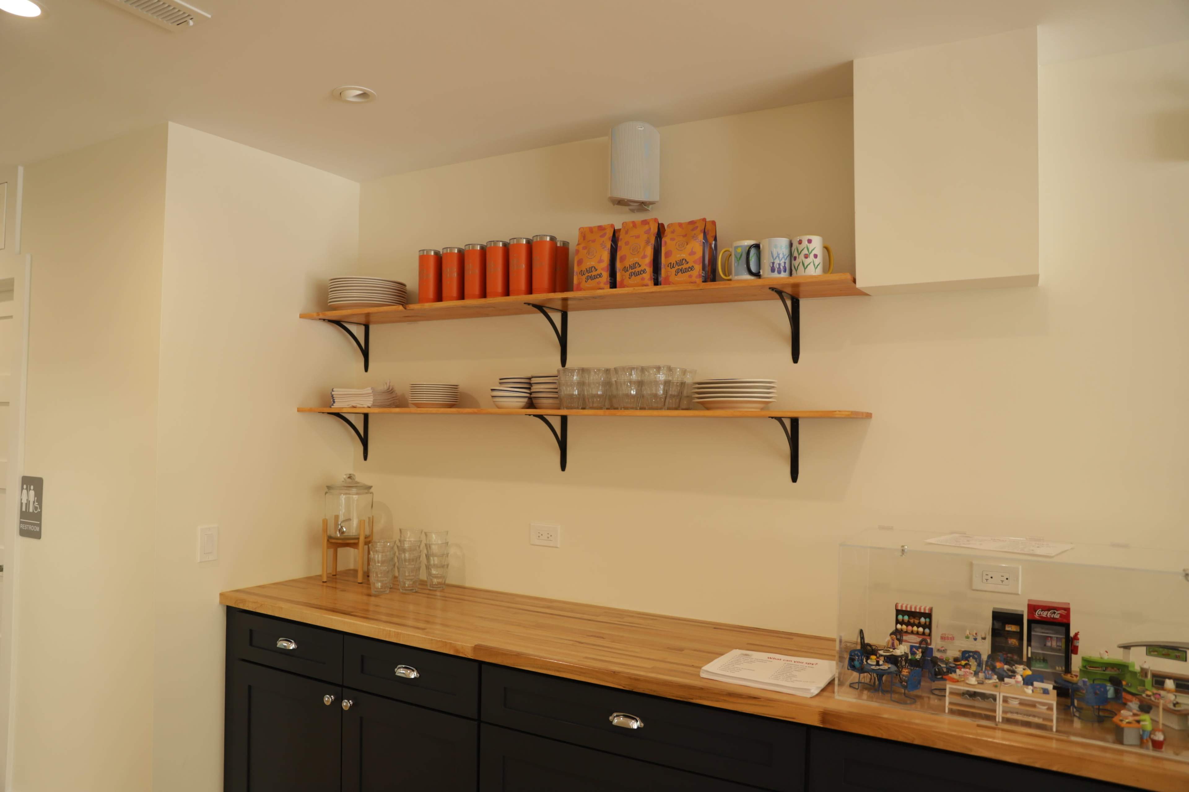 The image shows a kitchenette with wooden shelves displaying stacked plates, glassware, and orange juice containers above a countertop with a dark cabinet.