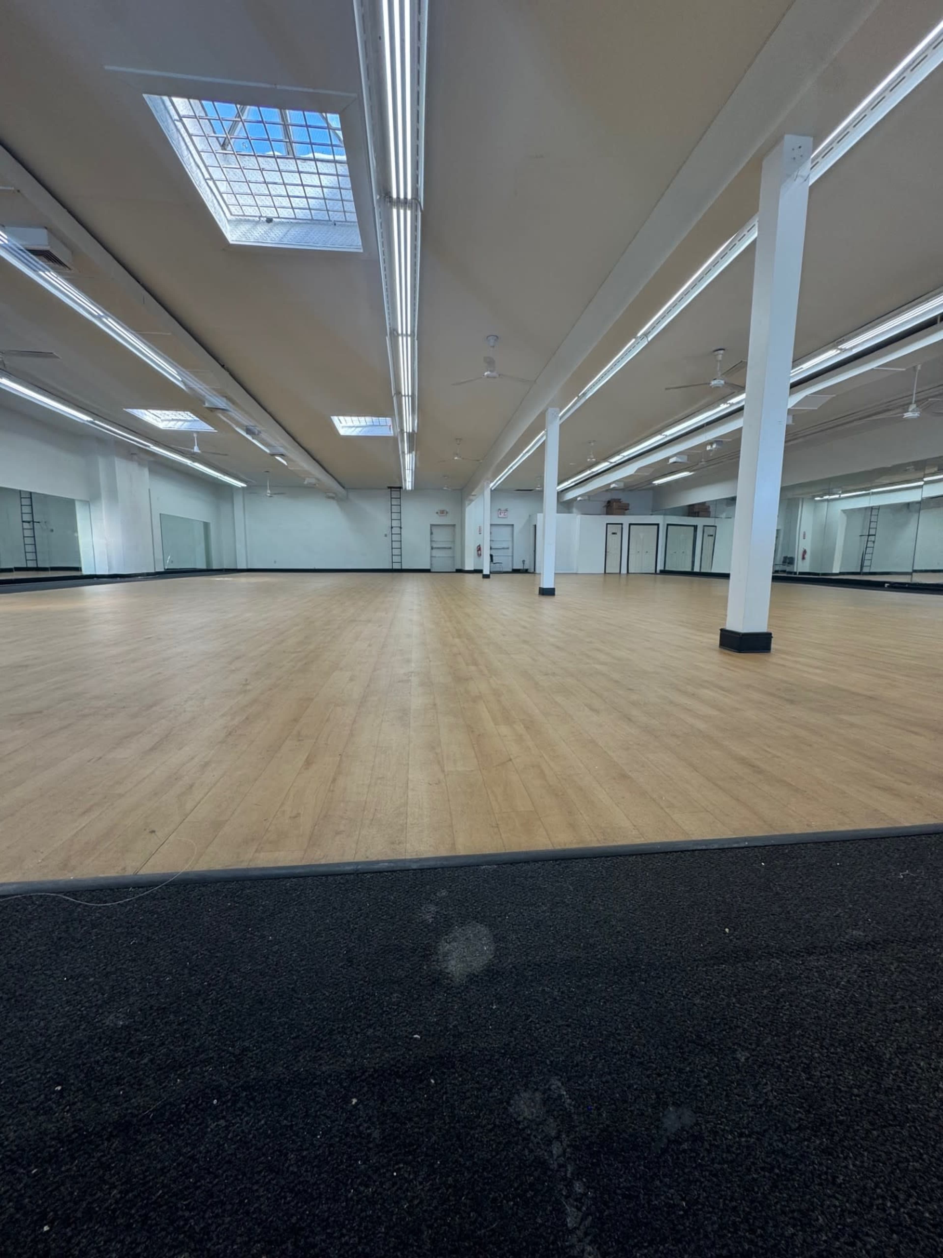 The image shows a spacious interior with a wooden floor, large mirrors along the walls, and ample overhead lighting.