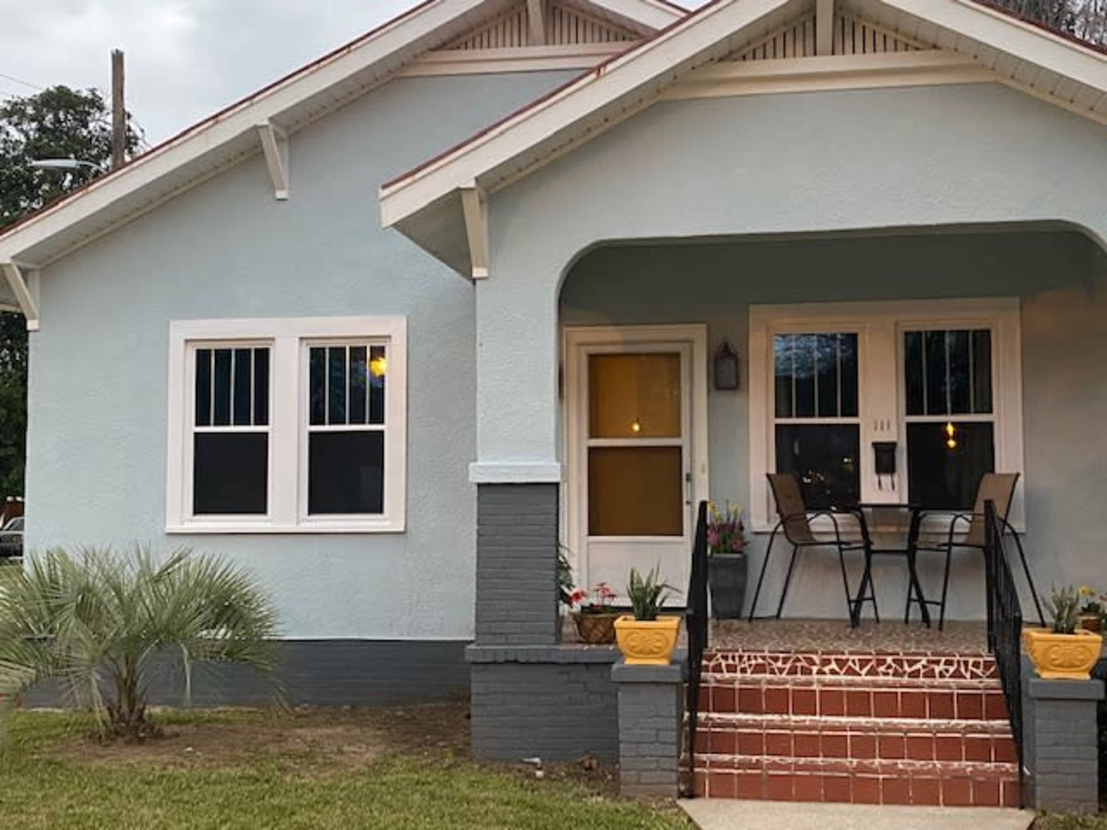 A light blue house with a covered front porch features two chairs and a small table, surrounded by well-maintained landscaping.