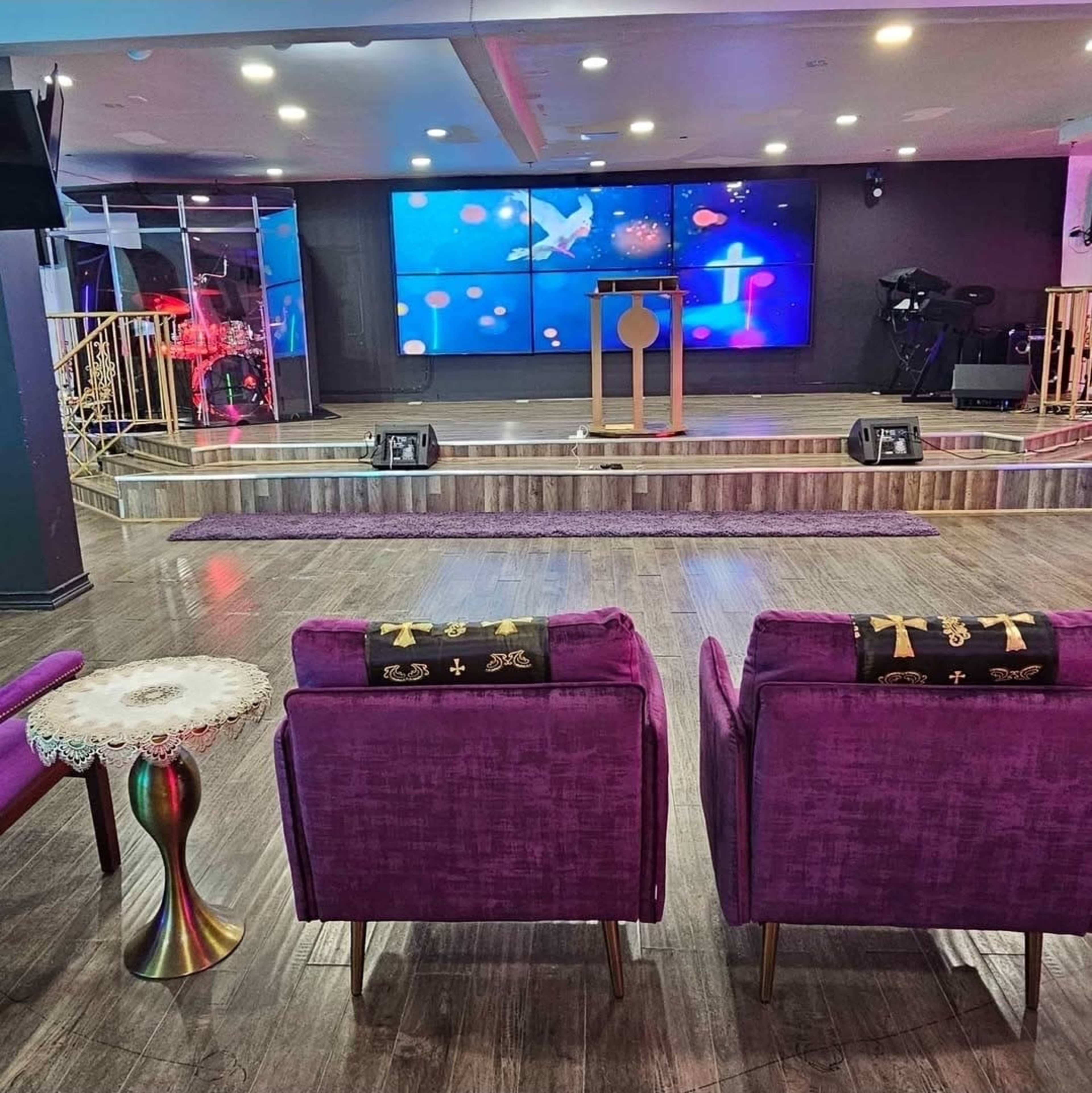 The image shows a modern worship space featuring two purple armchairs facing a stage with a large screen displaying colorful visuals.