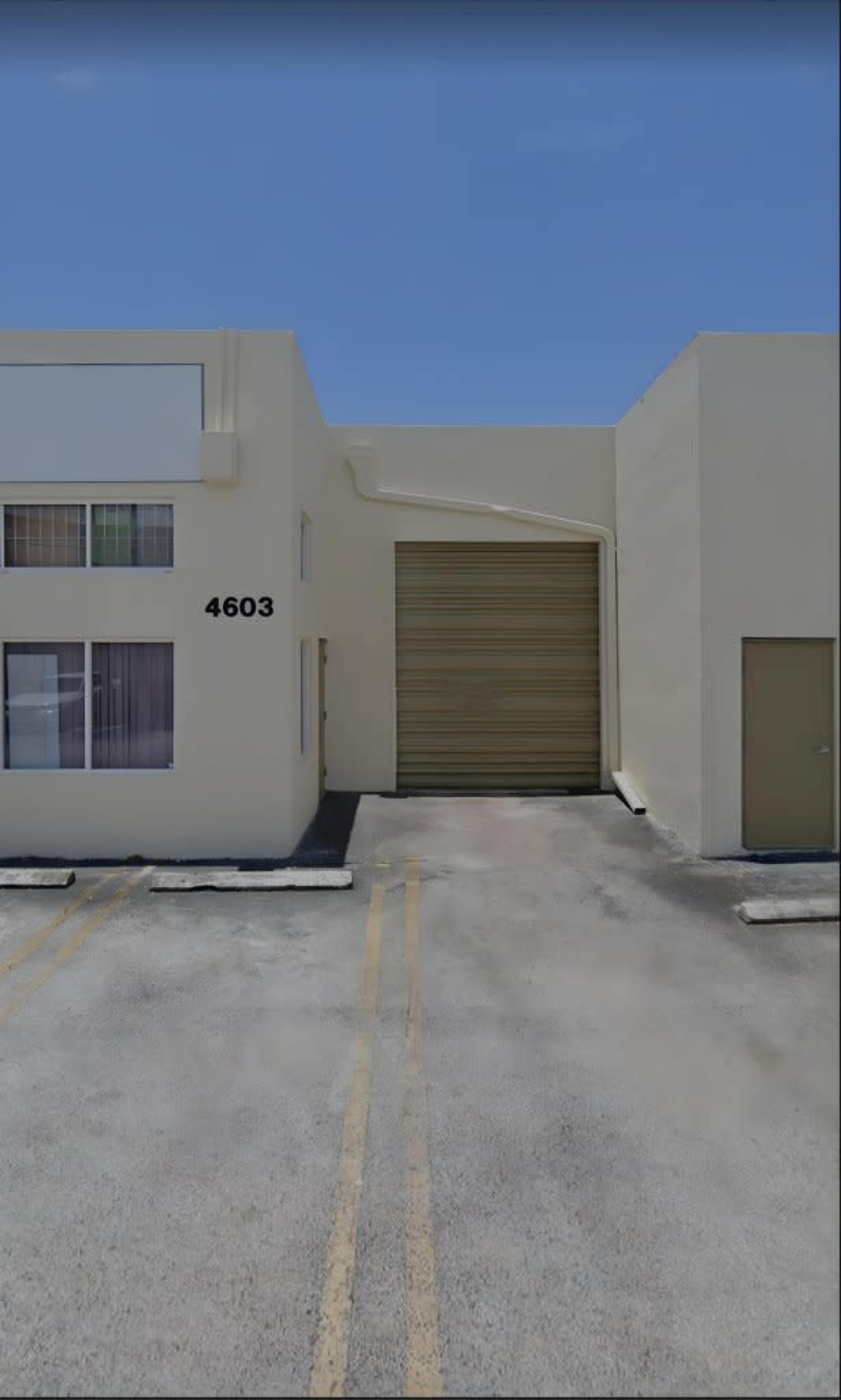 The image shows a light-colored commercial building with a large roll-up door and two windows, along with a parking area marked by yellow lines.