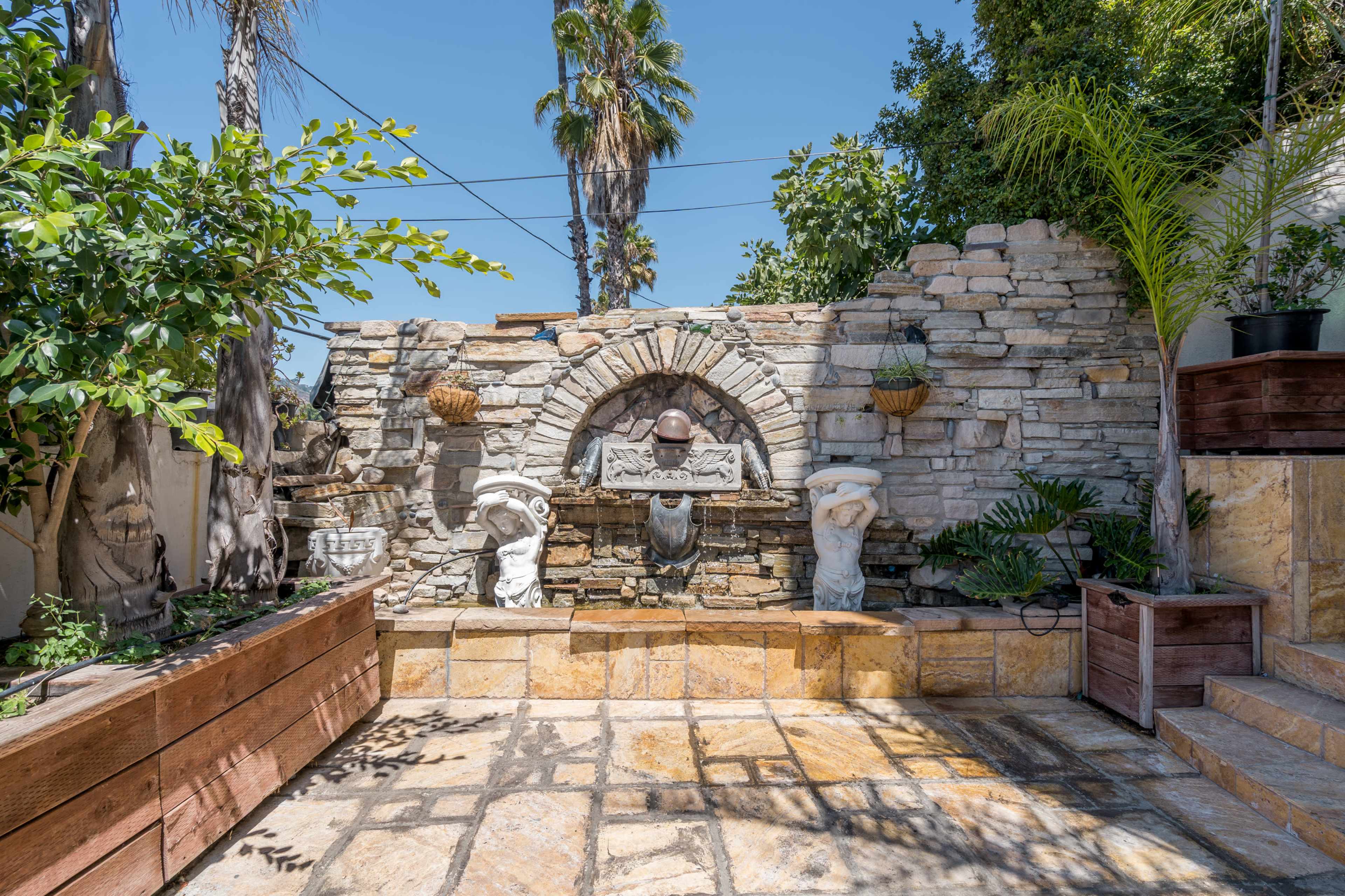 The image shows a landscaped outdoor space featuring a stone wall with a fountain, adorned with statues and surrounded by greenery and palm trees.