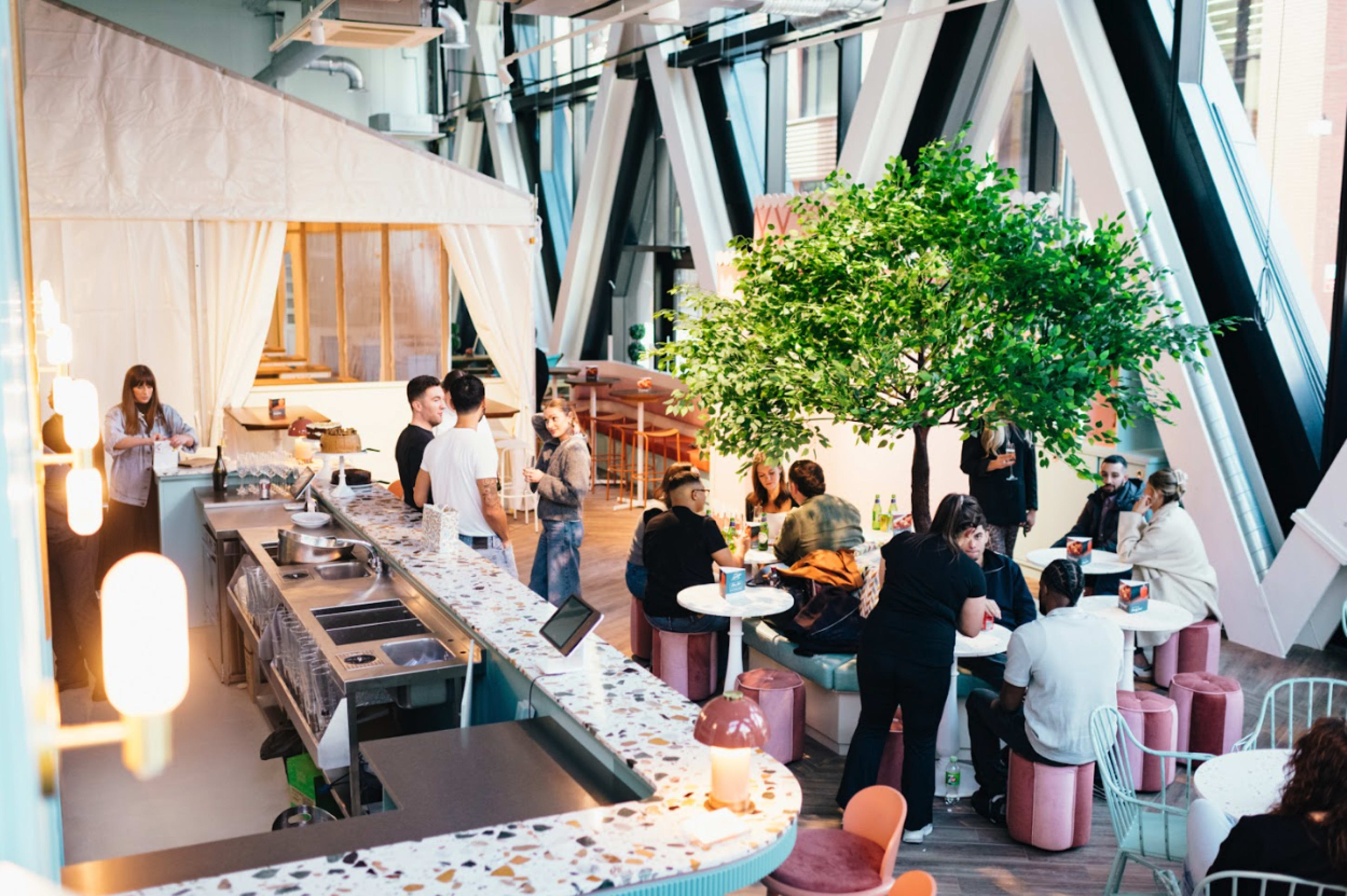 The image shows a modern café space with a large tree in the center, surrounded by several people seated at colorful stools and tables, while others gather near a kitchen area.