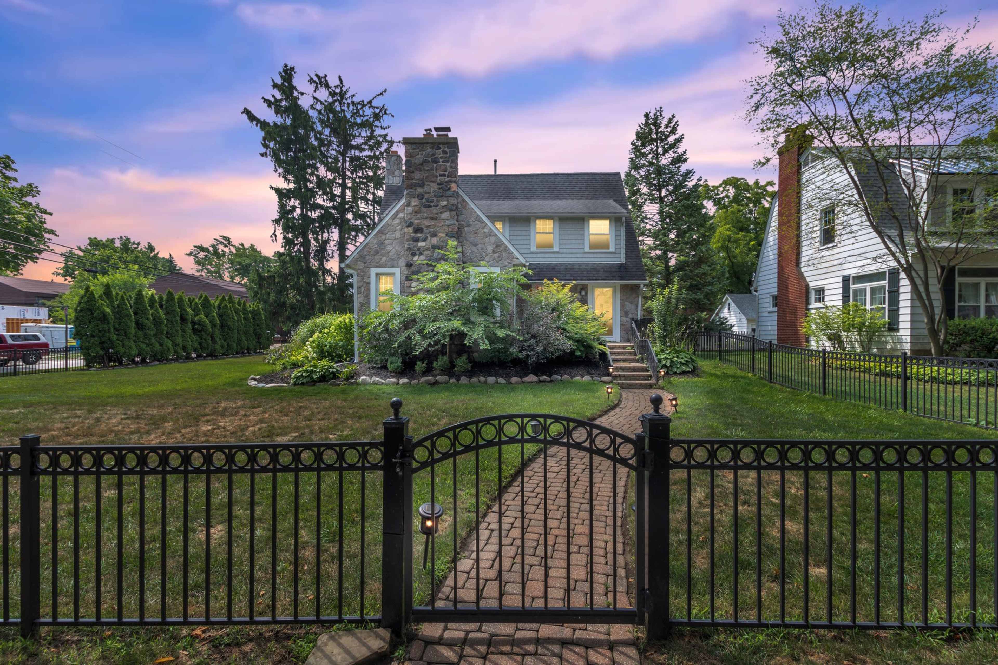 A stone house with a sloped roof and a front garden is enclosed by a black metal fence at dusk, with trees and neighboring houses in the background.