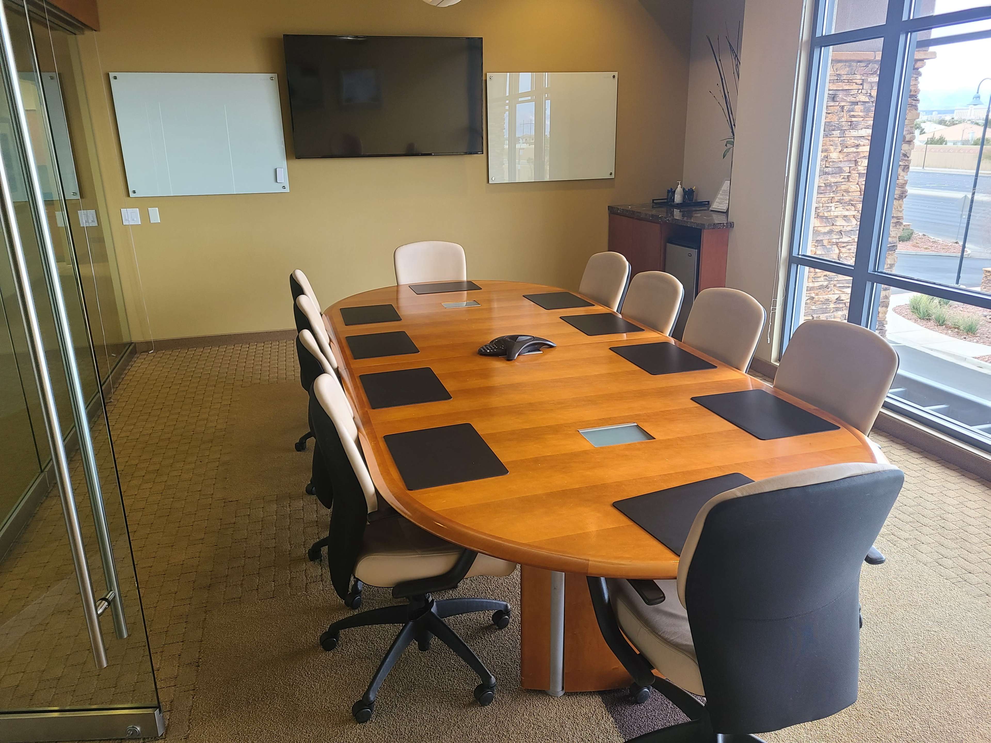 A conference room features a large oval wooden table surrounded by several office chairs, with a wall-mounted screen and whiteboards in the background.