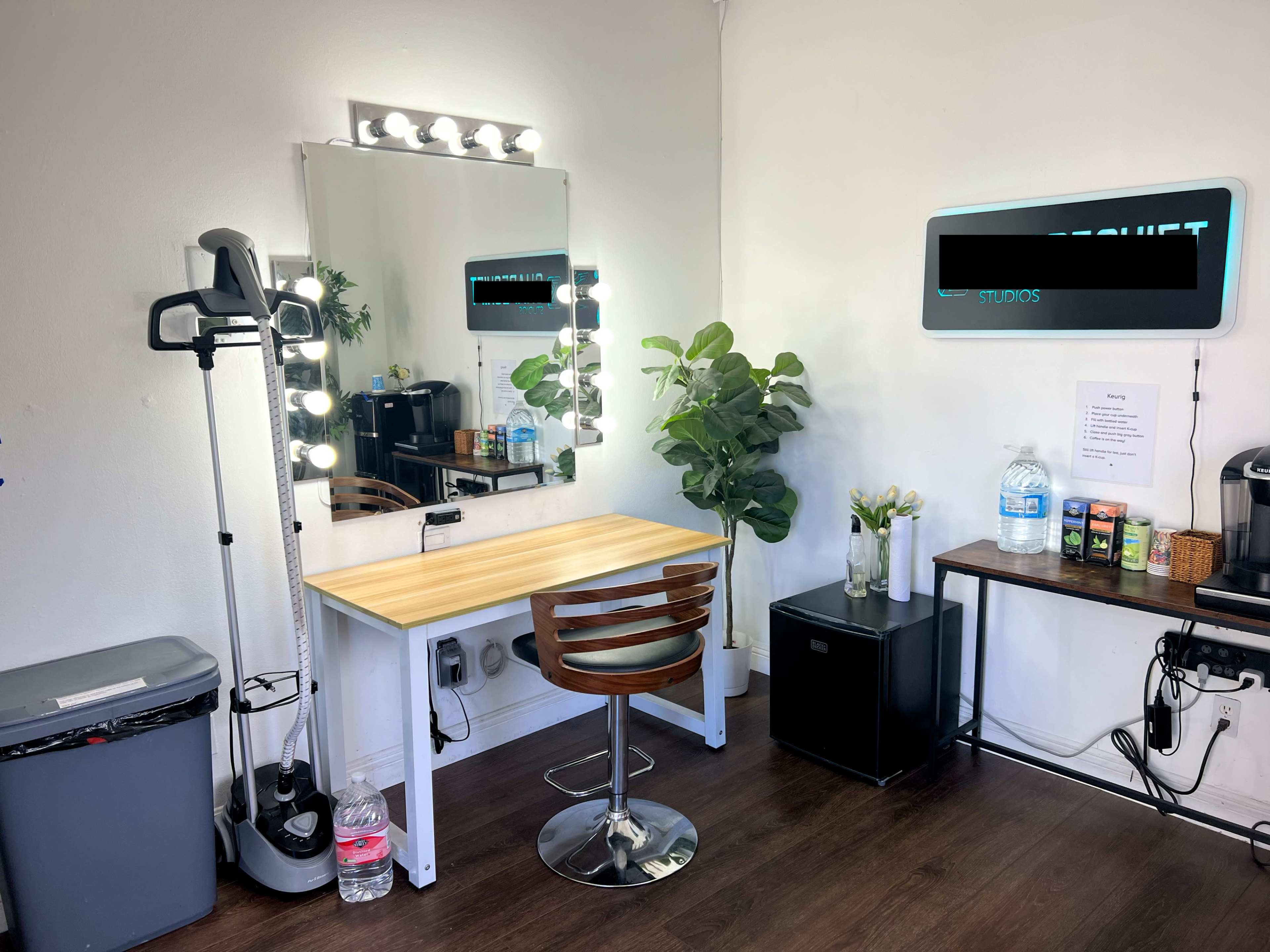 The image shows a well-lit vanity area with a mirror surrounded by bulbs, a wooden table with a chair, a garbage bin, and a small refrigerator, along with a plant and various items on a countertop.