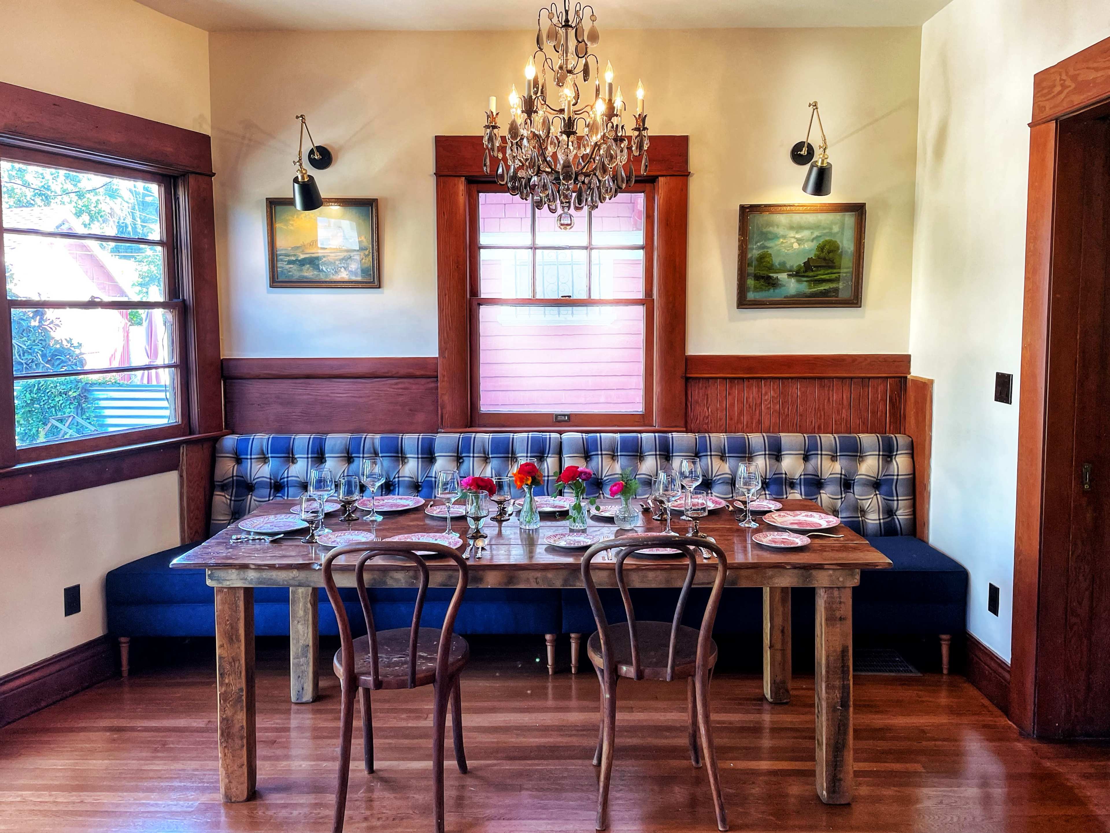 A dining room features a long wooden table set with plates and glasses, surrounded by wooden chairs, and a patterned banquette under framed paintings.