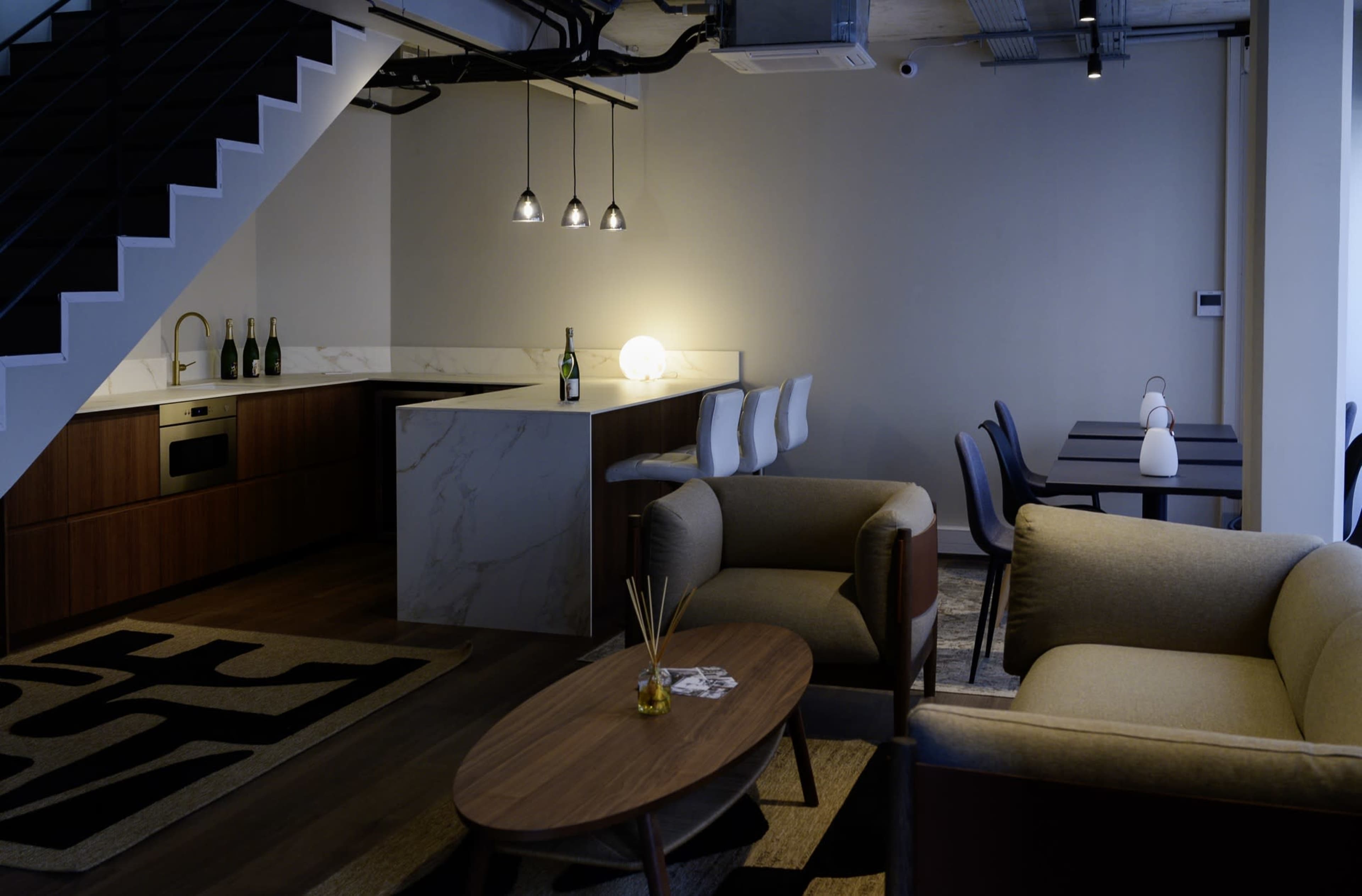 The image shows a modern interior of a lounge area featuring a kitchen with bar seating, a dining table, and a cozy seating arrangement.