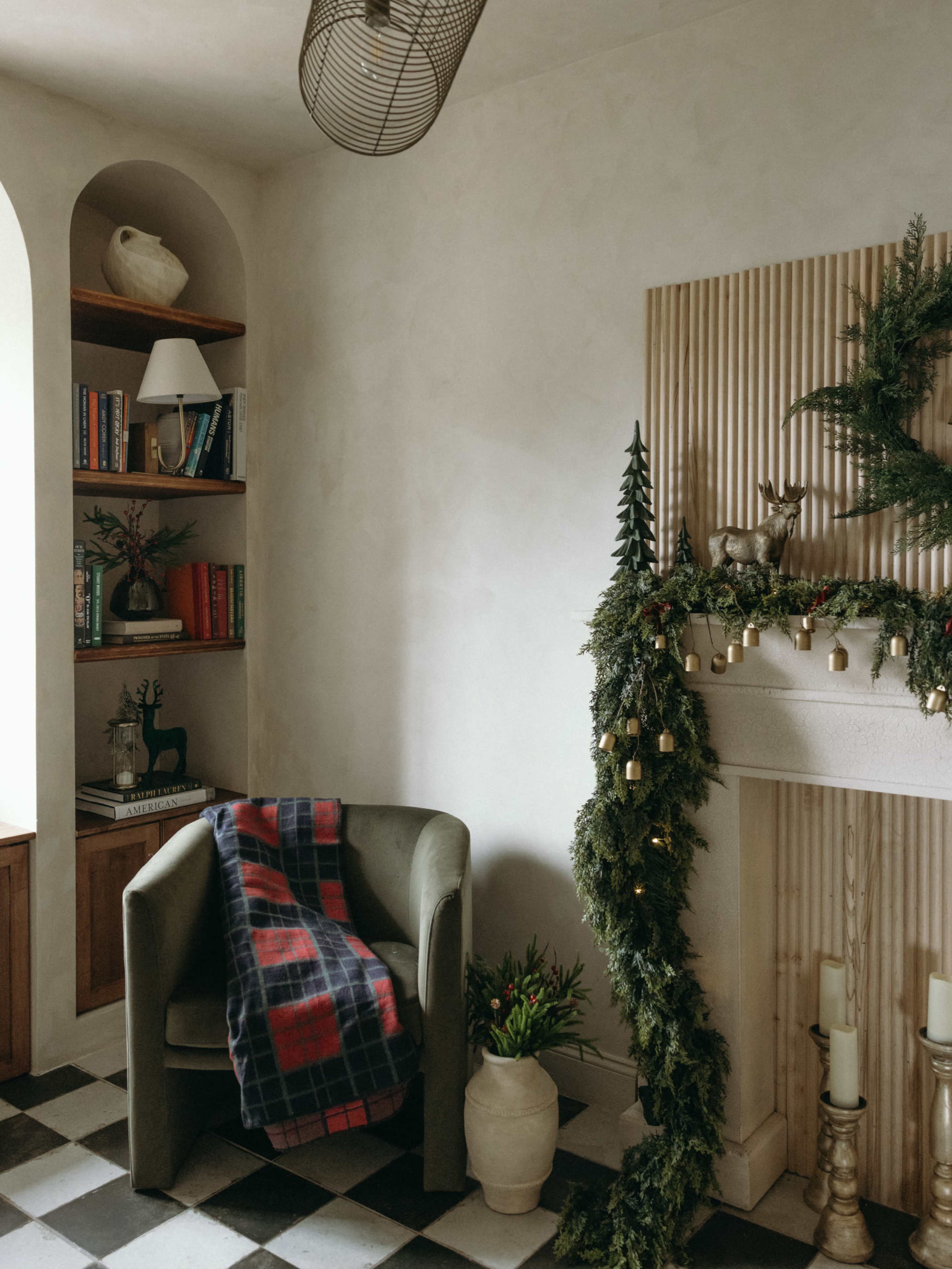 A cozy interior features a green armchair next to a bookshelf, a decorative fireplace adorned with garlands, and a plaid blanket draped over the chair.
