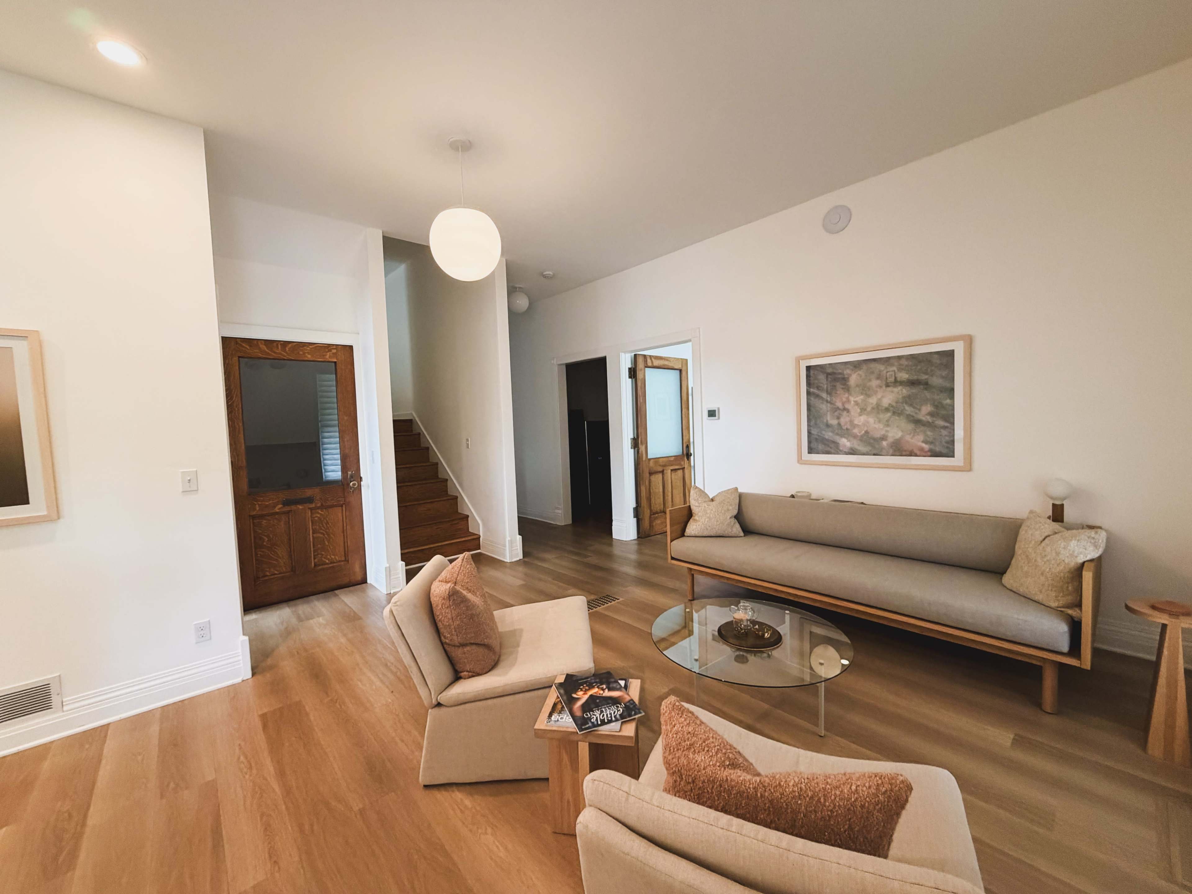 A modern living room features a light-colored sofa, two armchairs, a glass coffee table, and a doorway leading to a staircase.