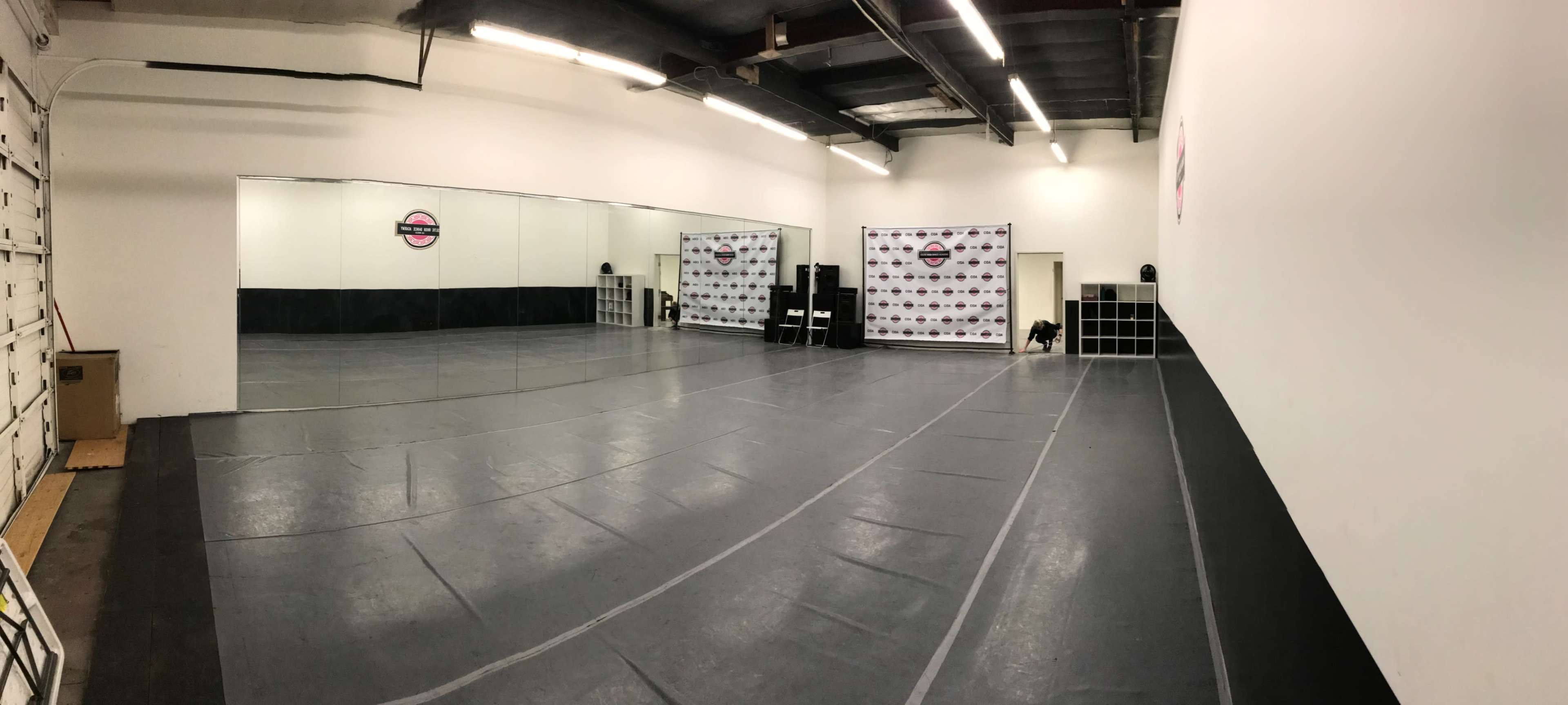 The image shows a spacious dance studio featuring a mirrored wall, a black and white backdrop, and a gray floor.