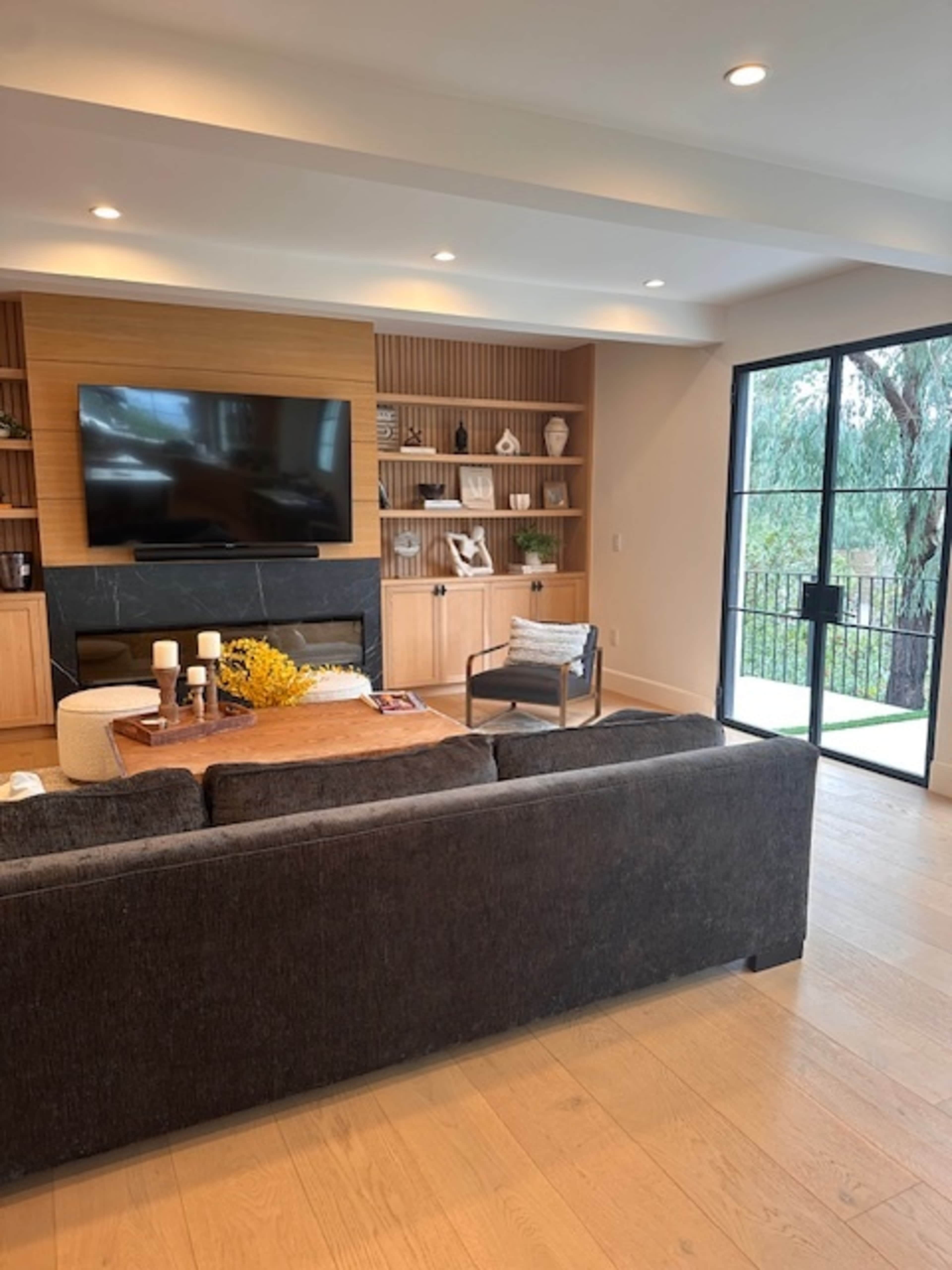 A modern living room features a dark sofa, a wooden coffee table with yellow flowers, a wall-mounted TV, and large windows overlooking greenery.