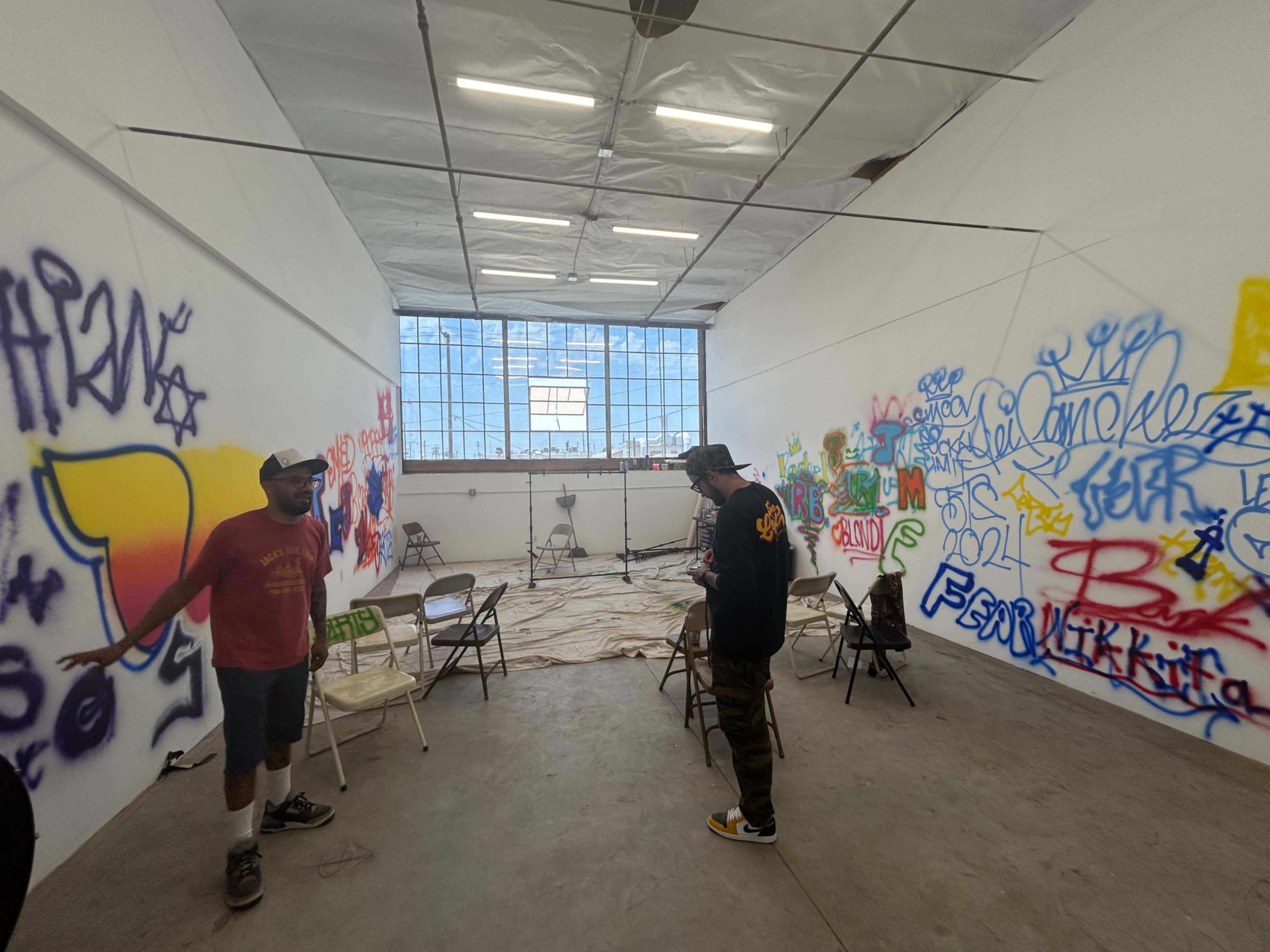 Two individuals stand in a room with graffiti-covered walls and chairs arranged on the floor, accompanied by a large window allowing natural light.