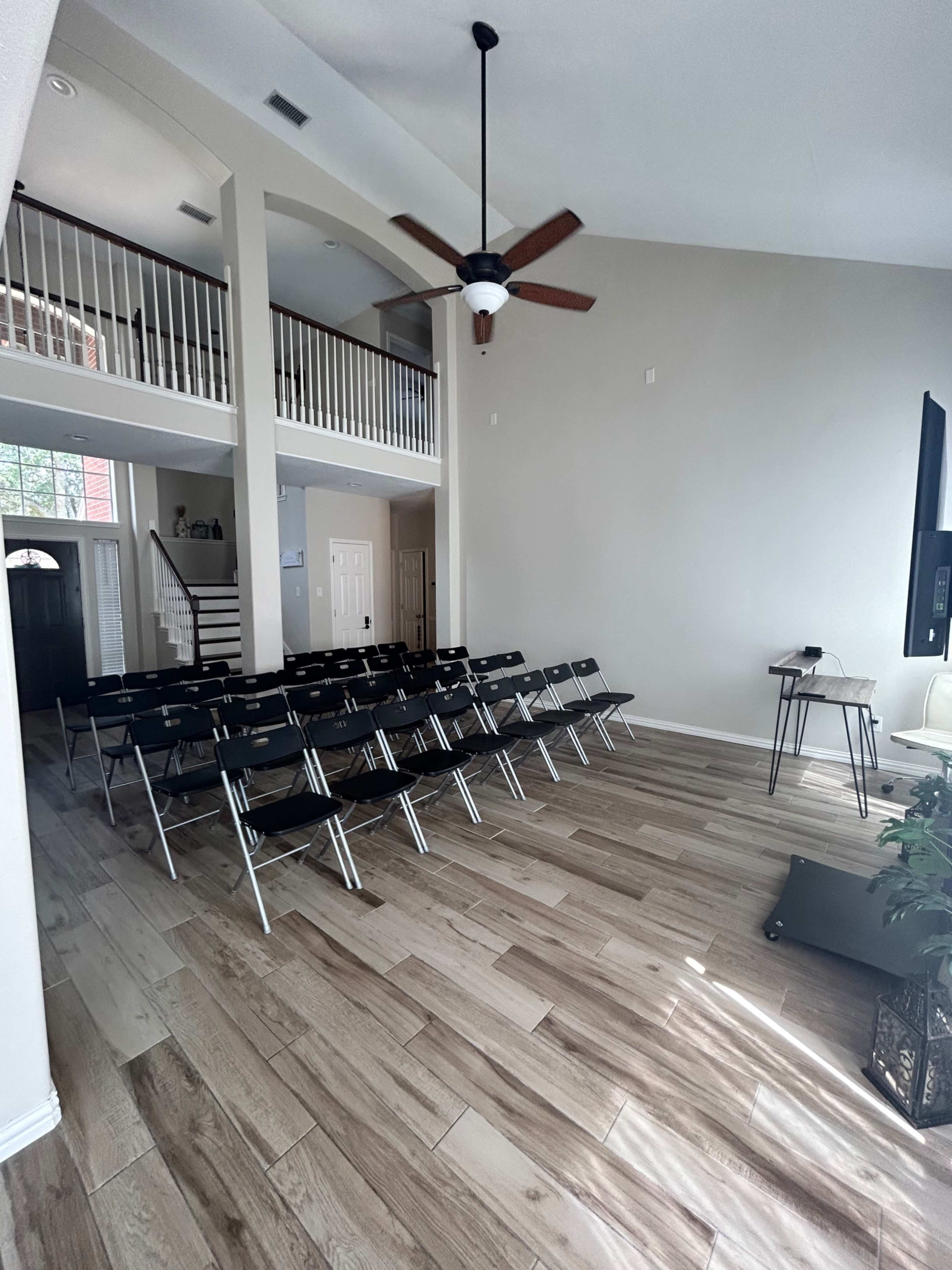 The image shows a spacious, light-colored room with rows of black folding chairs facing a television on a small stand, and a staircase leading to an upper level in the background.